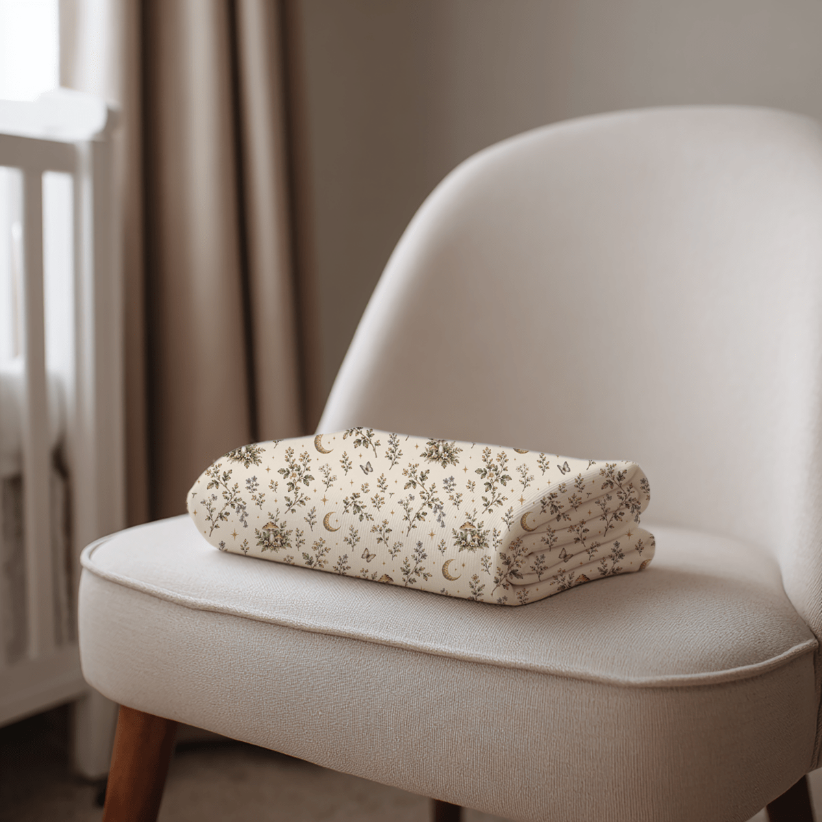 A folded, cream-colored Moonlit Sprigs Swaddle Blanket by Moon & Moss Nursery from Mirabilia Boutique, featuring a floral and crescent moon pattern, rests on a beige upholstered chair near a window and white crib.