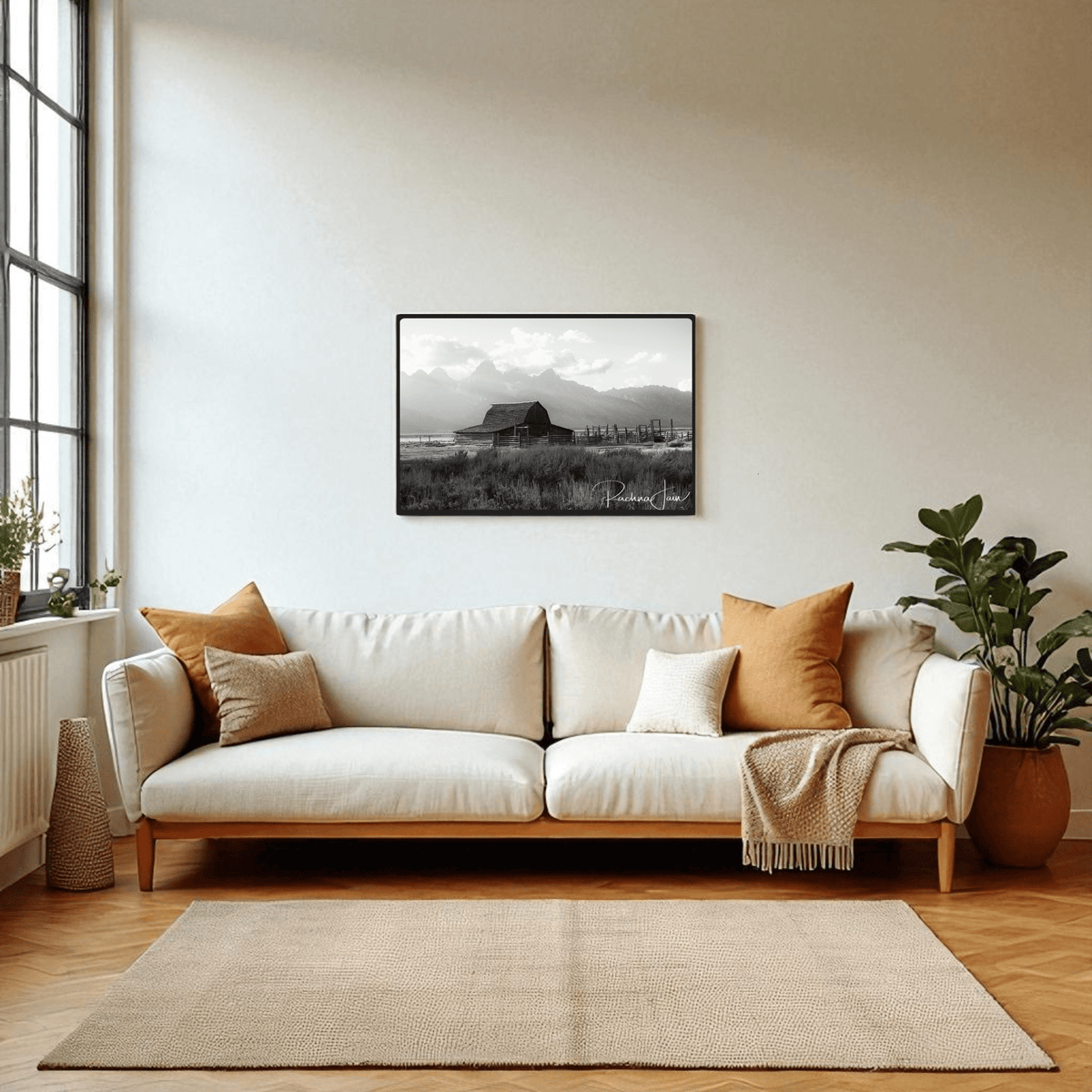A modern living room with a white sofa, earth-toned pillows, potted plants, and a large window features the "Moulton Barn Grand Teton" black and white framed art print by Rachna Jain from Mirabilia Boutique above the sofa.