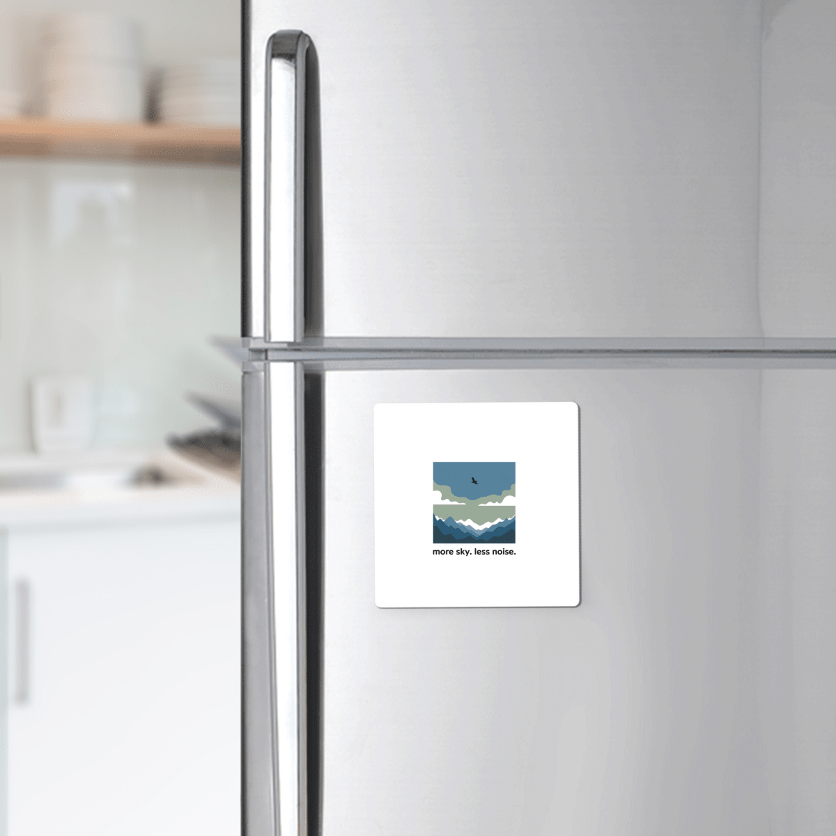 A silver fridge displays the “More Sky Less Noise Nature Magnet” by Mirabilia Boutique, featuring a minimalist landscape and bird. Ideal outdoor gift for minimalist decor lovers. Modern kitchen with white shelves in the background.