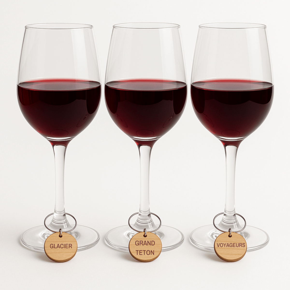 Three wine glasses with red wine, each featuring Mirabilia Boutique’s Autumn Lodge National Parks Wine Charms labeled “GLACIER,” “GRAND TETON,” and “VOYAGEURS,” are arranged side by side on a white surface.