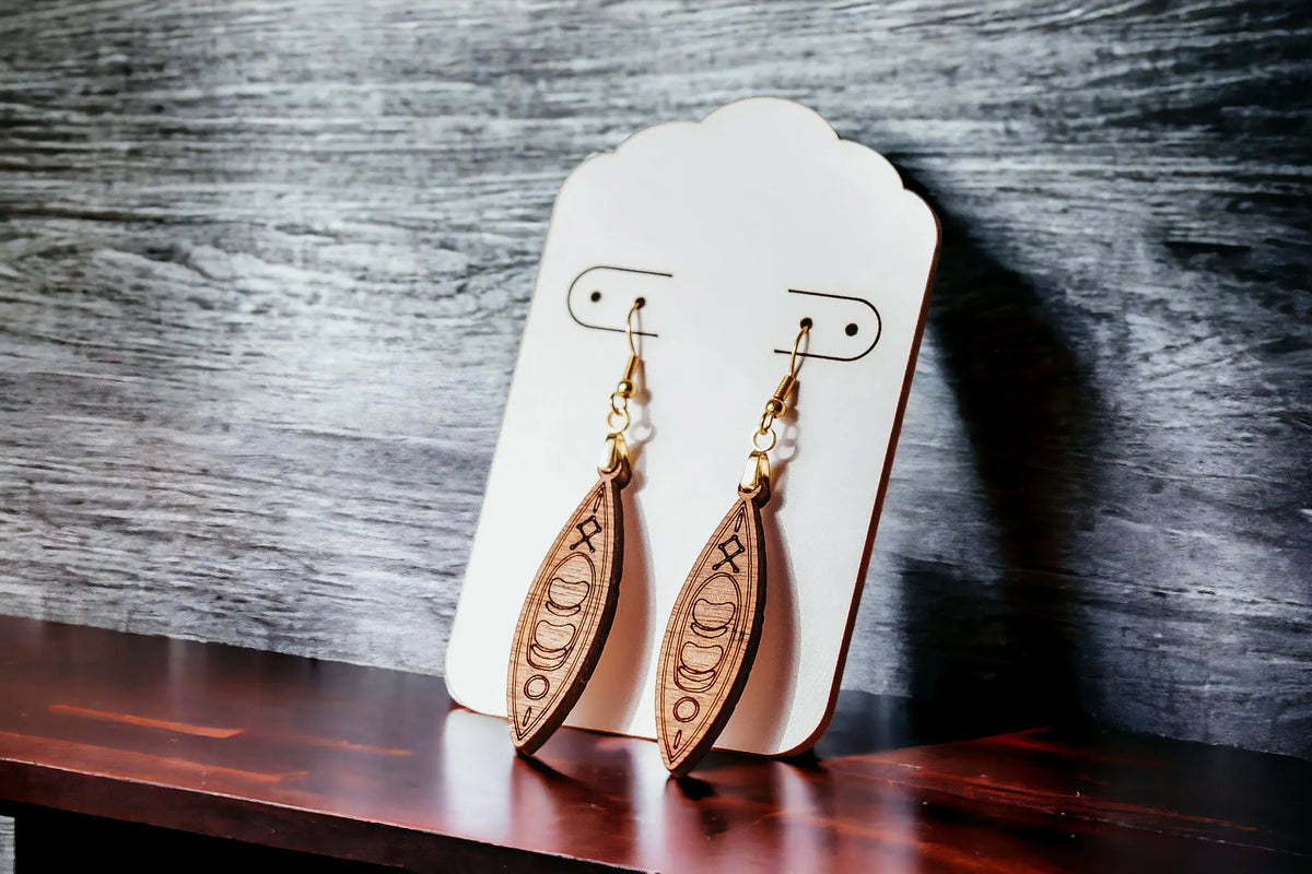 Kayak Earrings by Mirabilia Boutique—laser-cut wooden dangle earrings with intricate designs for outdoor lovers—are displayed on a white card against a gray background and dark wood, making them the perfect canoe gift for river girls and paddlers.