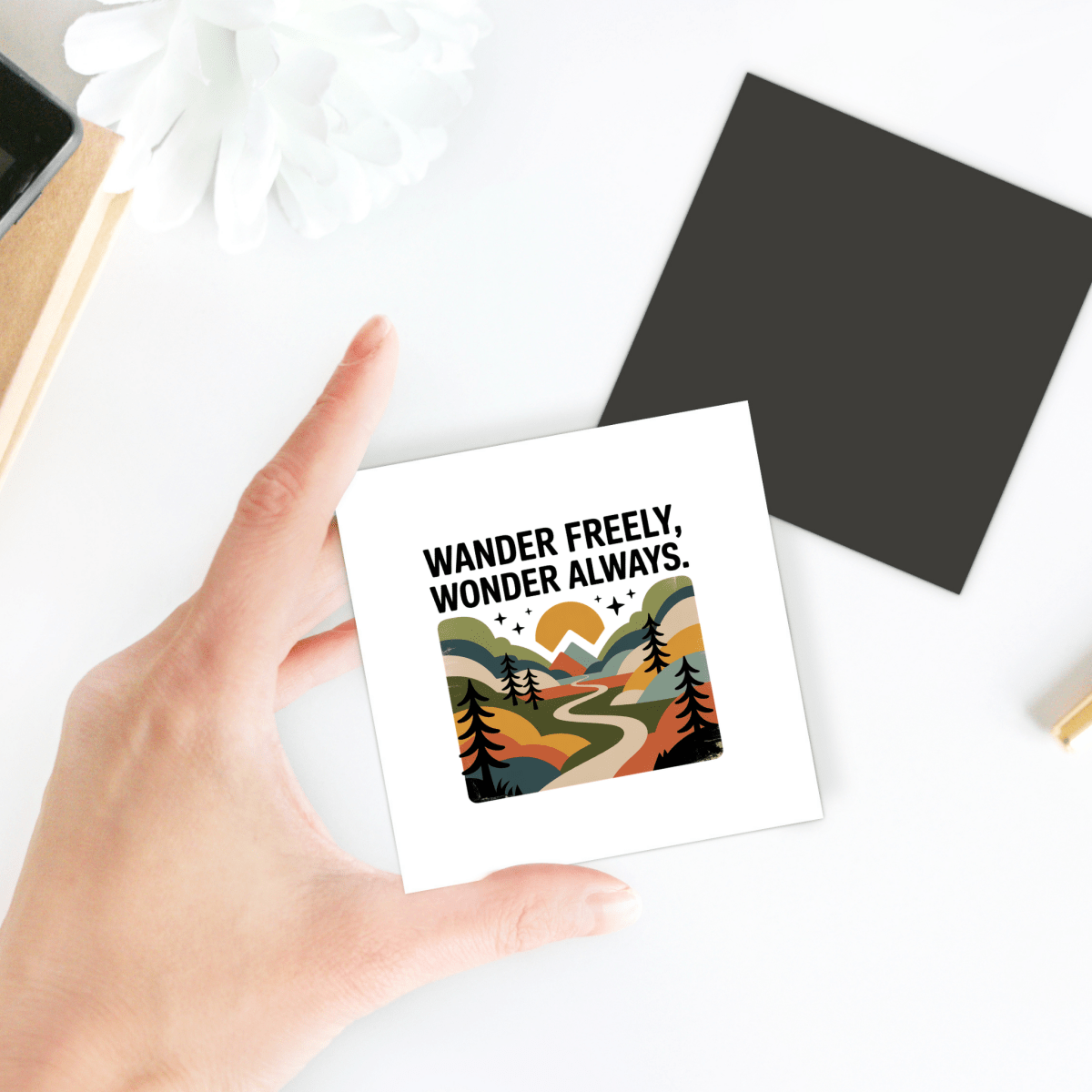 A hand holds the Mirabilia Boutique "Wander Freely Wonder Always Adventure Magnet," a square magnet with colorful mountains, trees, and a winding path. The card says, “WANDER FREELY, WONDER ALWAYS.” Great outdoorsy gift!.