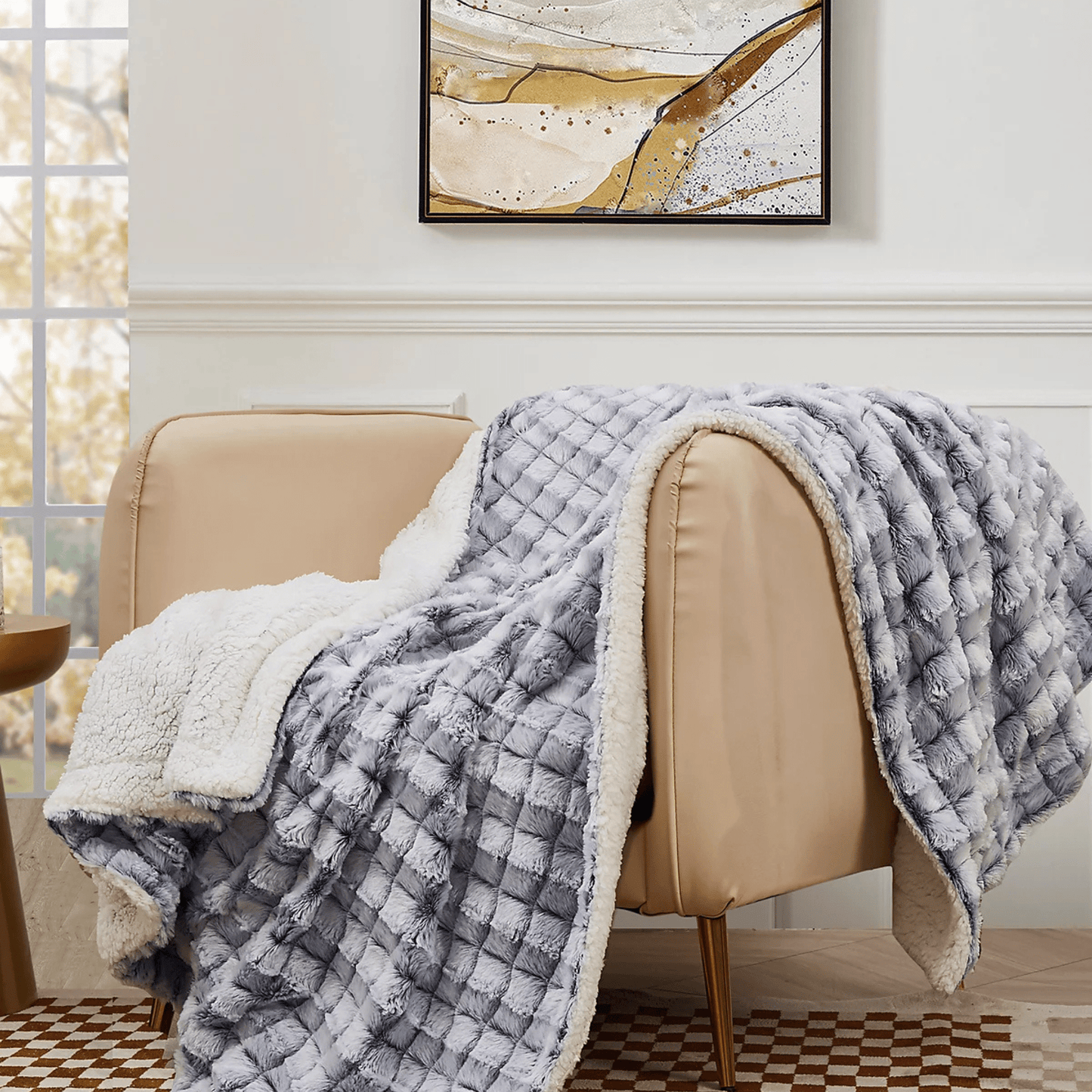 The Mirabilia Boutique Custom Fall Throw Blanket, a plush gray and white checkered cozy autumn gift, is draped over a beige armchair in a room with modern abstract art and a large window—perfect for adding warmth to your home.