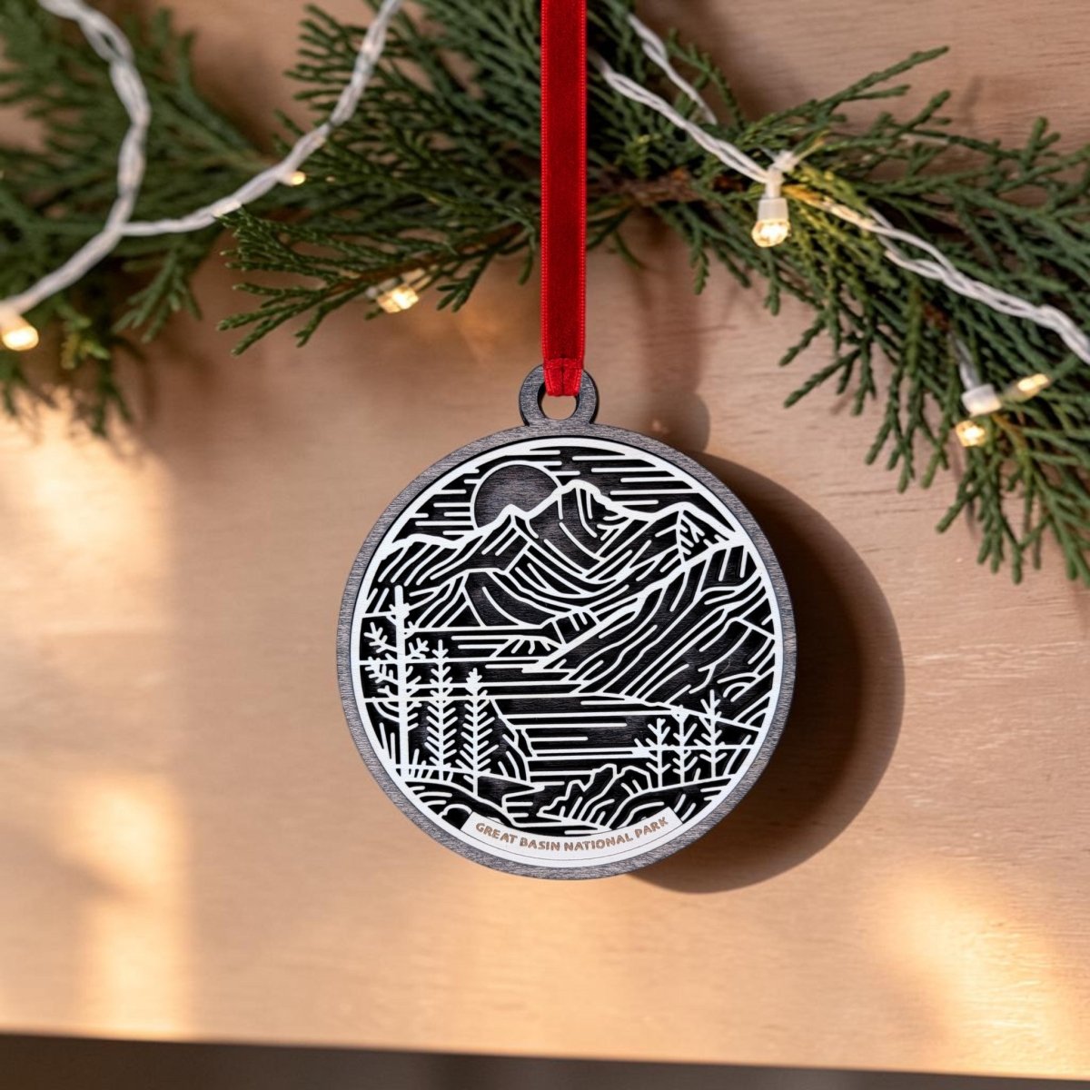 A black and white line art ornament from Mirabilia Boutique, featuring a mountain and forest design, hangs from a red ribbon. Greenery and string lights in the background highlight the unique beauty of this Great Basin National Park Line Art Ornament. This wooden keepsake is labeled “Great Basin National Park.”.