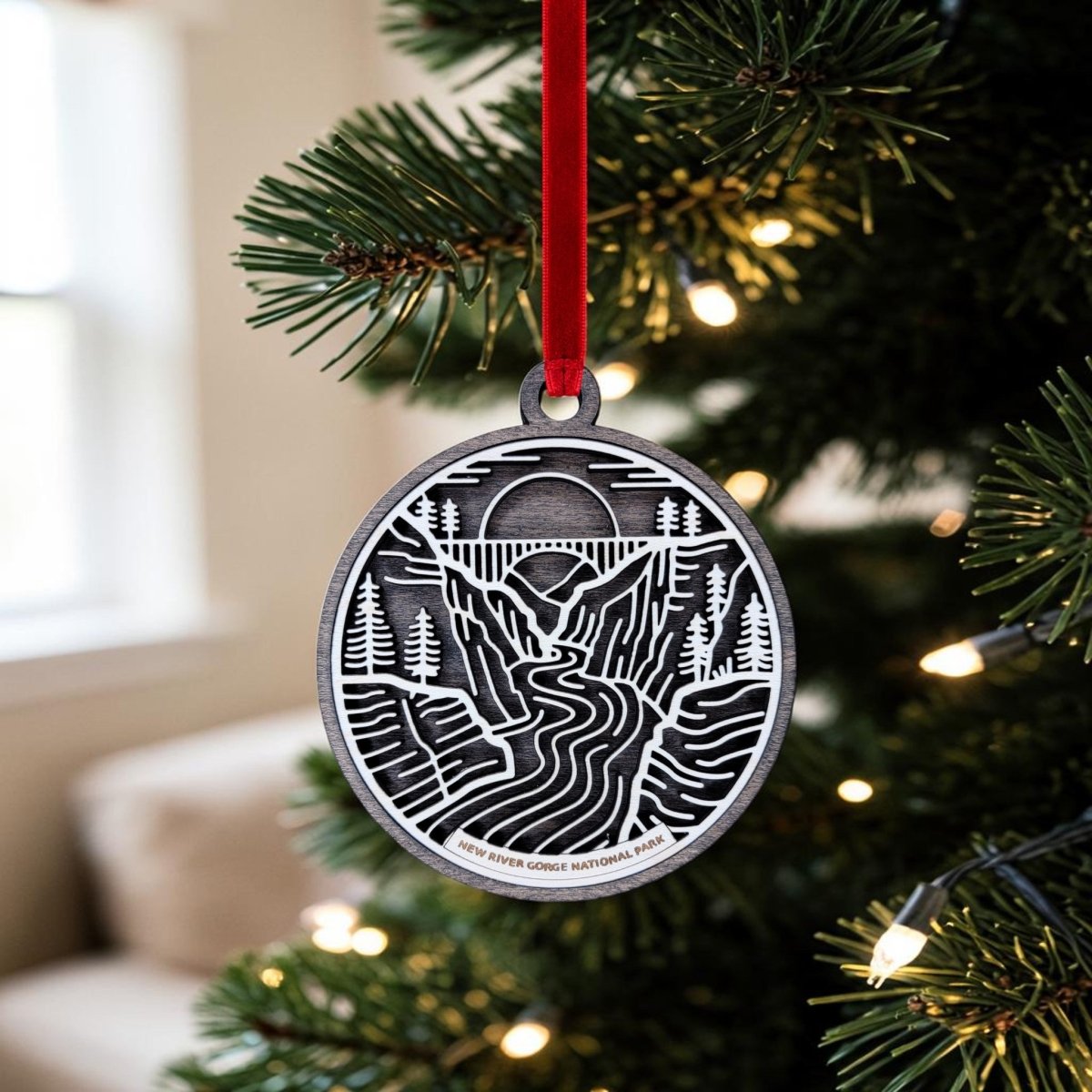 A New River Gorge National Park Line Art Christmas Ornament by Mirabilia Boutique hangs from a red ribbon on a Christmas tree with lights. This metal ornament features the New River Gorge Bridge spanning a river, framed by trees, mountains, and a sunset—capturing the beauty of New River Gorge National Park.