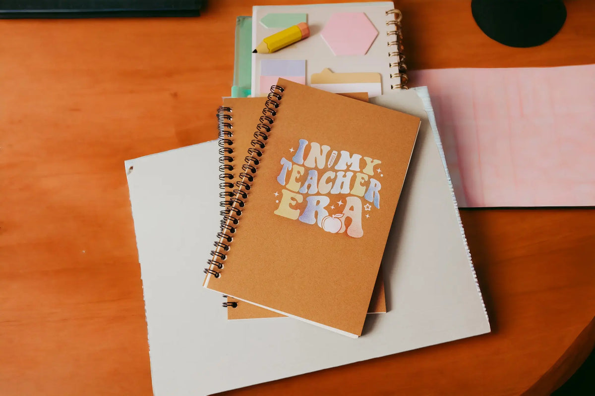 The Mirabilia Boutique "In My Teacher Era Notebook"—an aesthetic spiral journal ideal as a back-to-school teacher gift—rests on a wooden desk with pink paper and colorful sticky notes, making it a unique addition to any teacher's supplies.