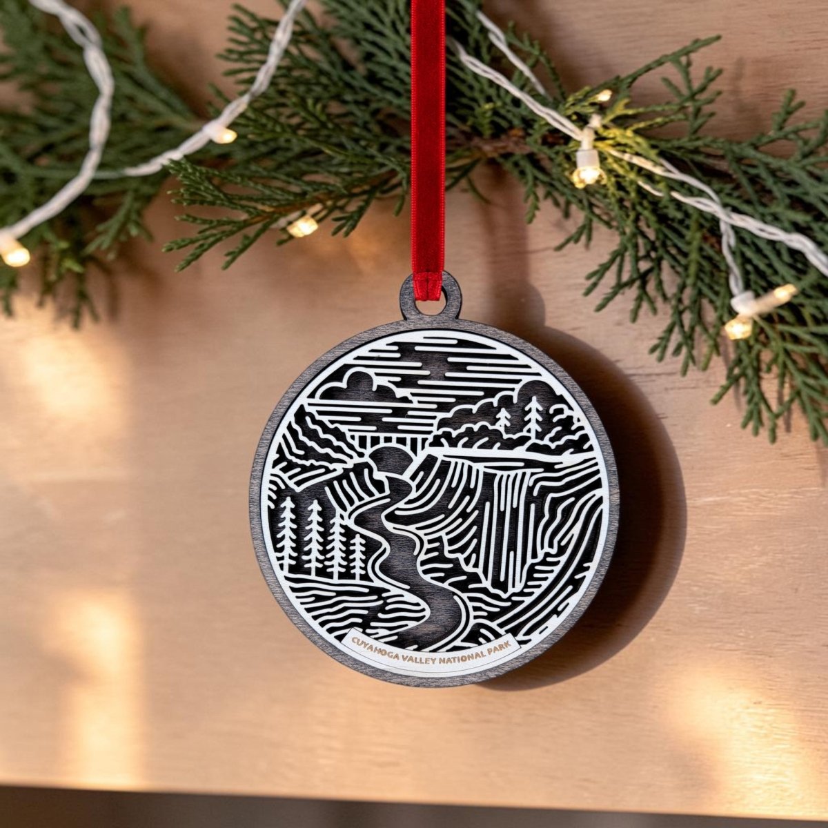 The Cuyahoga Valley National Park Christmas Ornament by Mirabilia Boutique features an oval wooden shape with a black and white landscape illustration of Brandywine Falls. It hangs from a red ribbon, displayed against a backdrop of evergreen branches and string lights, and is inscribed with "Cuyahoga Valley National Park.