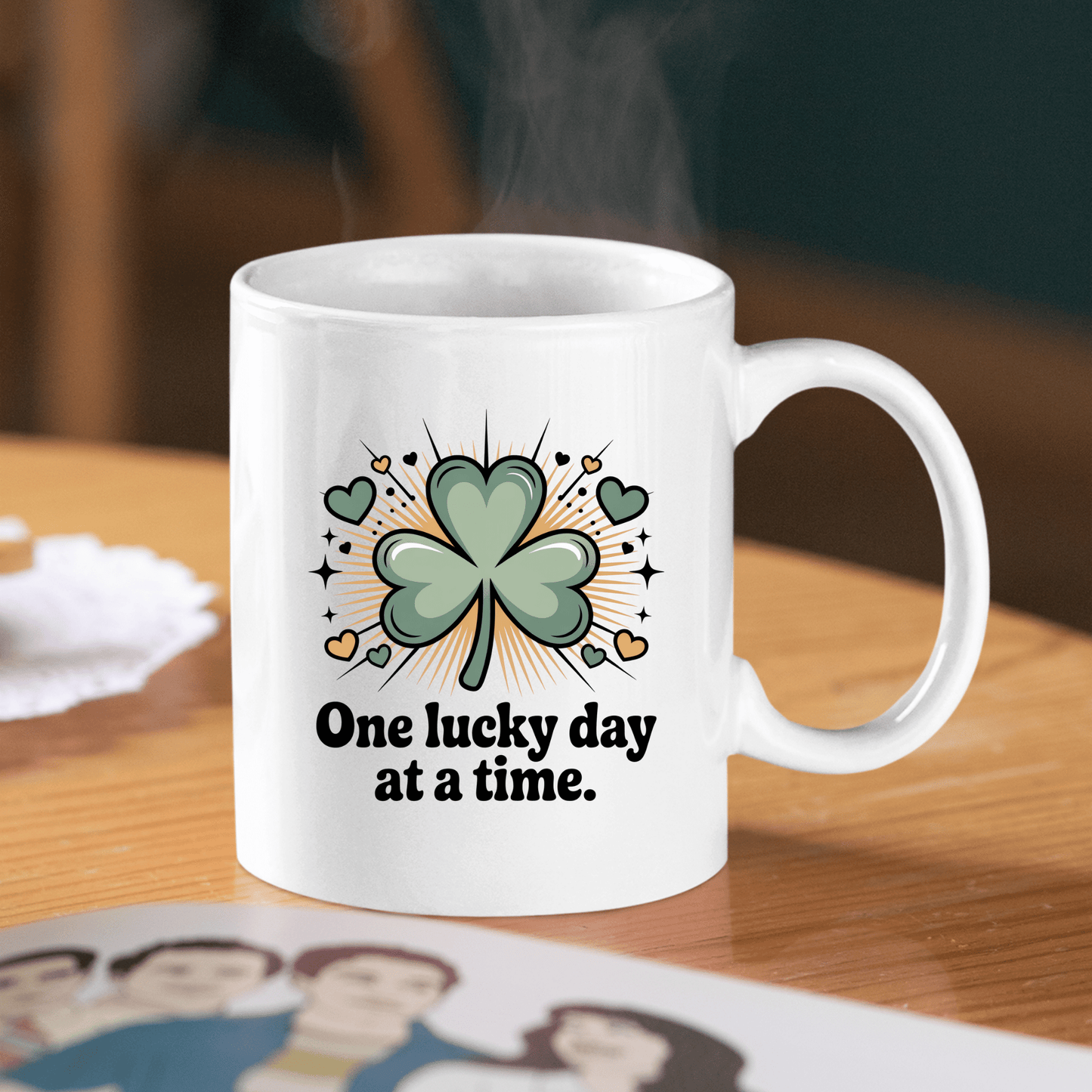 The Mirabilia Boutique One Lucky Day at a Time Mug – Shamrock Ceramic Cup sits on a wooden table, displaying a sparkling green shamrock and hearts with the uplifting phrase, “One lucky day at a time.” Steam wafts from the cup.