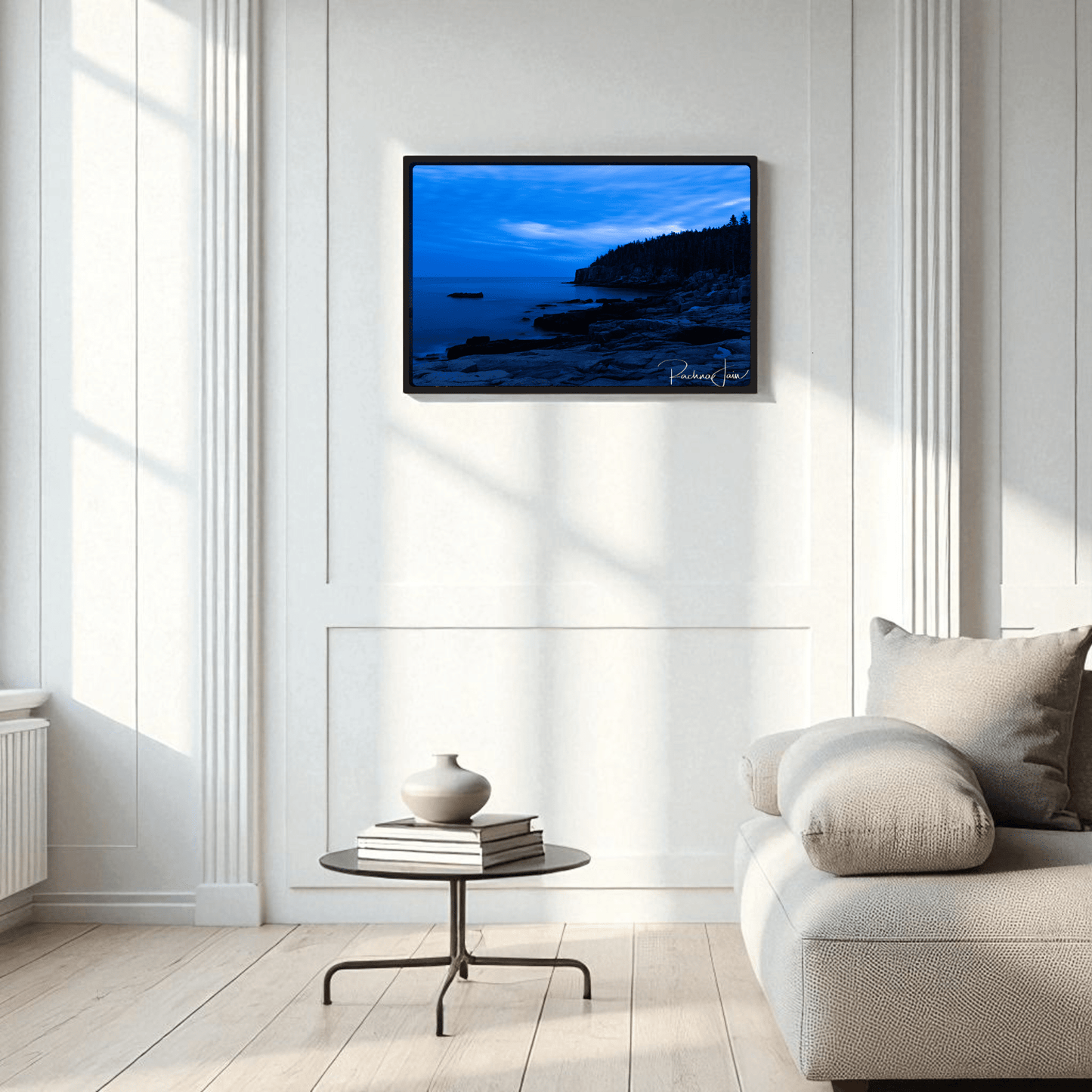 A modern living room with white paneled walls, a light gray sofa, and a round black coffee table displays Mirabilia Boutique’s Otter Point Acadia | Blue Hour Framed Art Print—coastal photography as striking wall art.