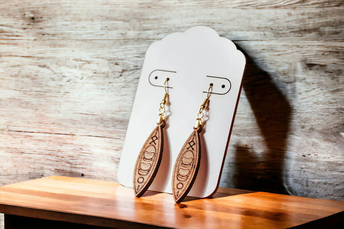 A pair of Kayak Earrings by Mirabilia Boutique—laser cut wooden dangle earrings for outdoor lovers, river girls, and paddlers—hang from a white display card on a light wooden surface. Perfect unique accessories for adventure enthusiasts.