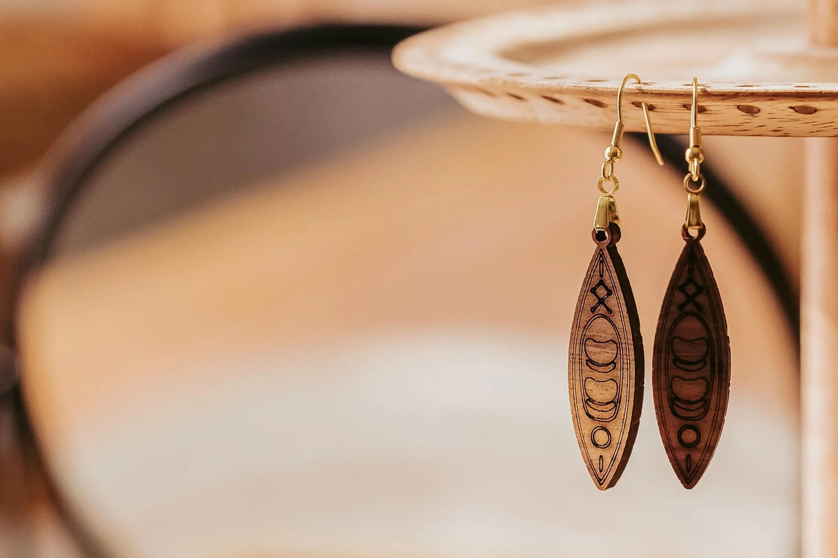 Mirabilia Boutique's Kayak Earrings are laser-cut wooden dangles featuring canoe designs. Perfect for outdoor lovers and paddlers, these lightweight earrings are displayed on a circular wooden stand with a softly blurred, warm-toned background.