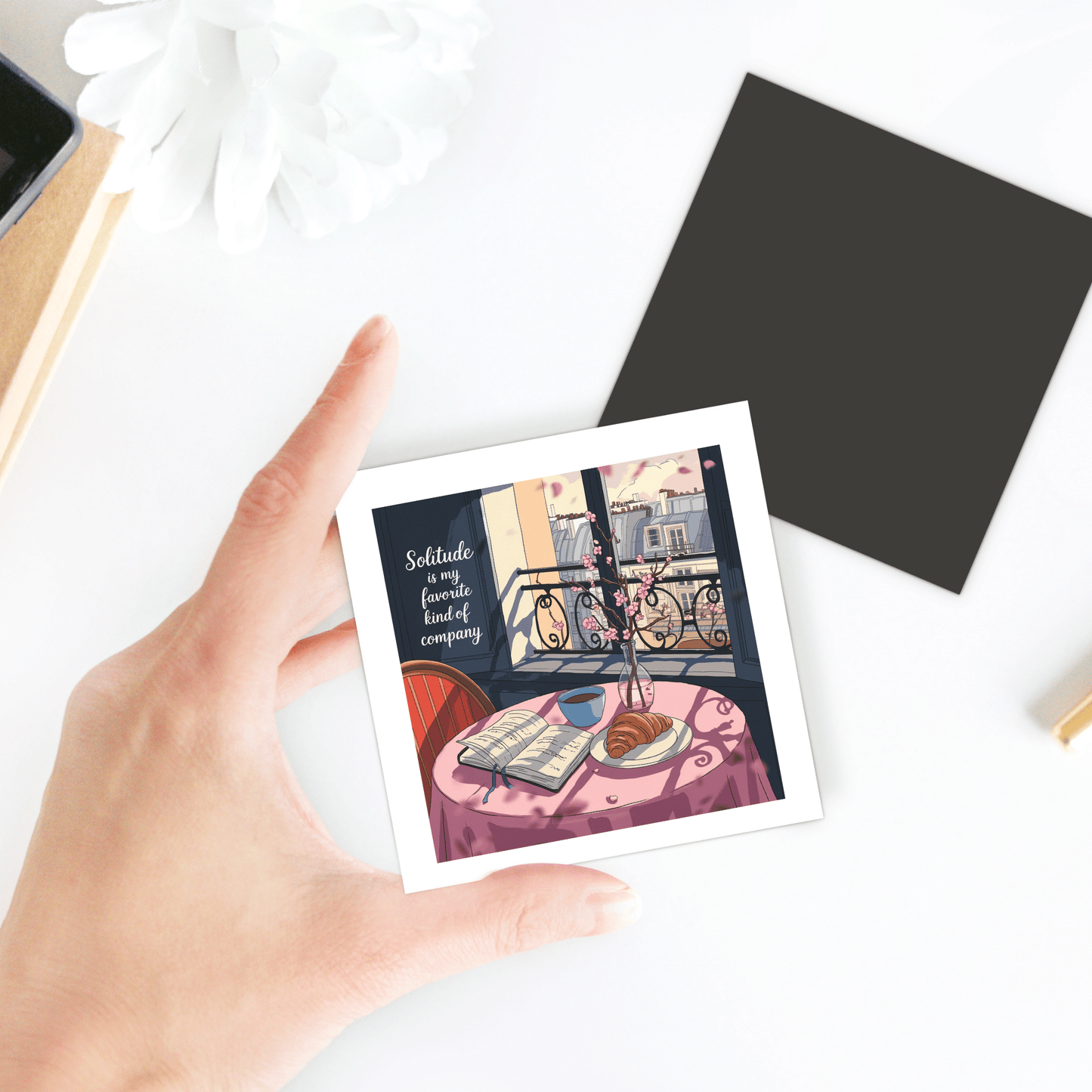 A hand holds the Solitude Magnet – Paris Morning Still Life by Mirabilia Boutique, featuring a cozy table scene with book, coffee, croissant, and city view—Parisian apartment decor. The magnet reads: "Solitude is my favorite kind of company.