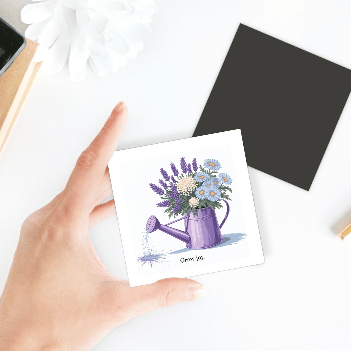 A hand holds the Mirabilia Boutique "Grow Joy Magnet – Watering Can & Soft Garden Florals," showing a purple can with lavender and white flowers. Perfect for gardeners or floral kitchen decor, with a black square and white flower in the background.
