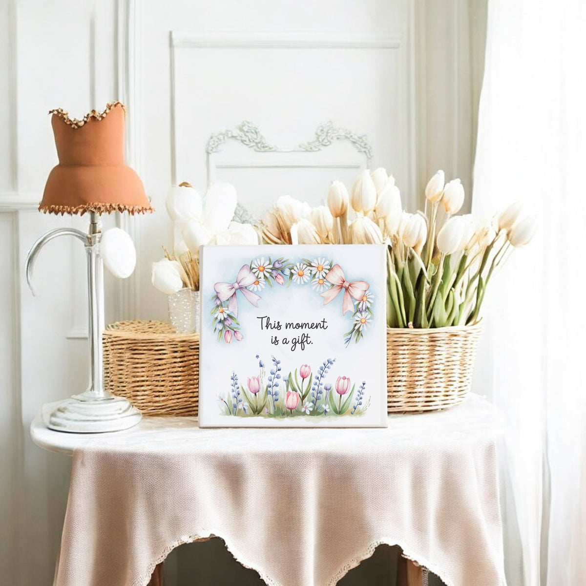 The Mirabilia Boutique "This Moment Is a Gift Canvas" (8x8 spring floral art) is displayed on a beige-draped table with white tulips in wicker baskets, a vintage lamp, and a white-paneled wall for a cozy slow living decor vibe.