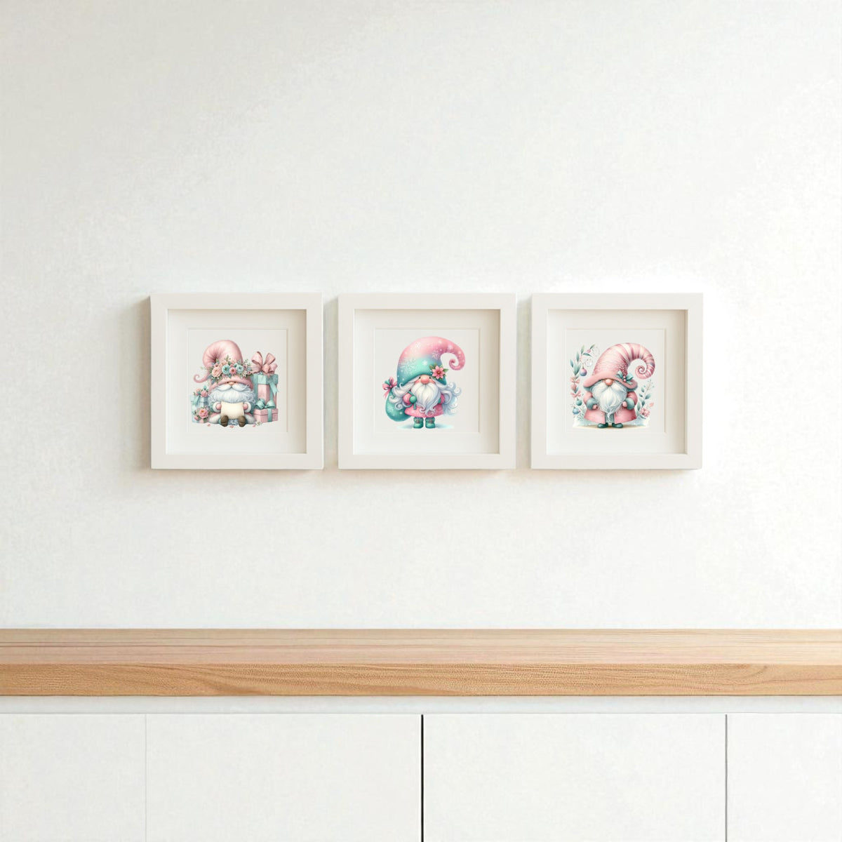 The Pastel Gnome Wall Art Trio by Mirabilia Boutique features three framed prints with pink-hatted gnomes on a clean white wall above a light wood cabinet—perfect for whimsical nursery or winter holiday decor.
