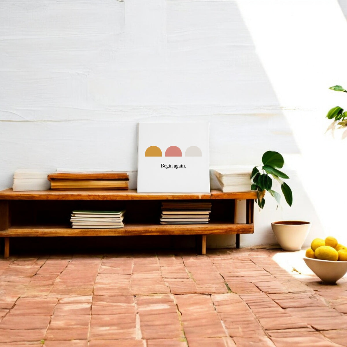 A low wooden bench with stacked books and the Mirabilia Boutique "Begin Again Canvas – Soft Summer Affirmation Wall Art" rests against a white wall. Nearby, a potted plant and a bowl of lemons sit on terracotta tiles in sunlight.