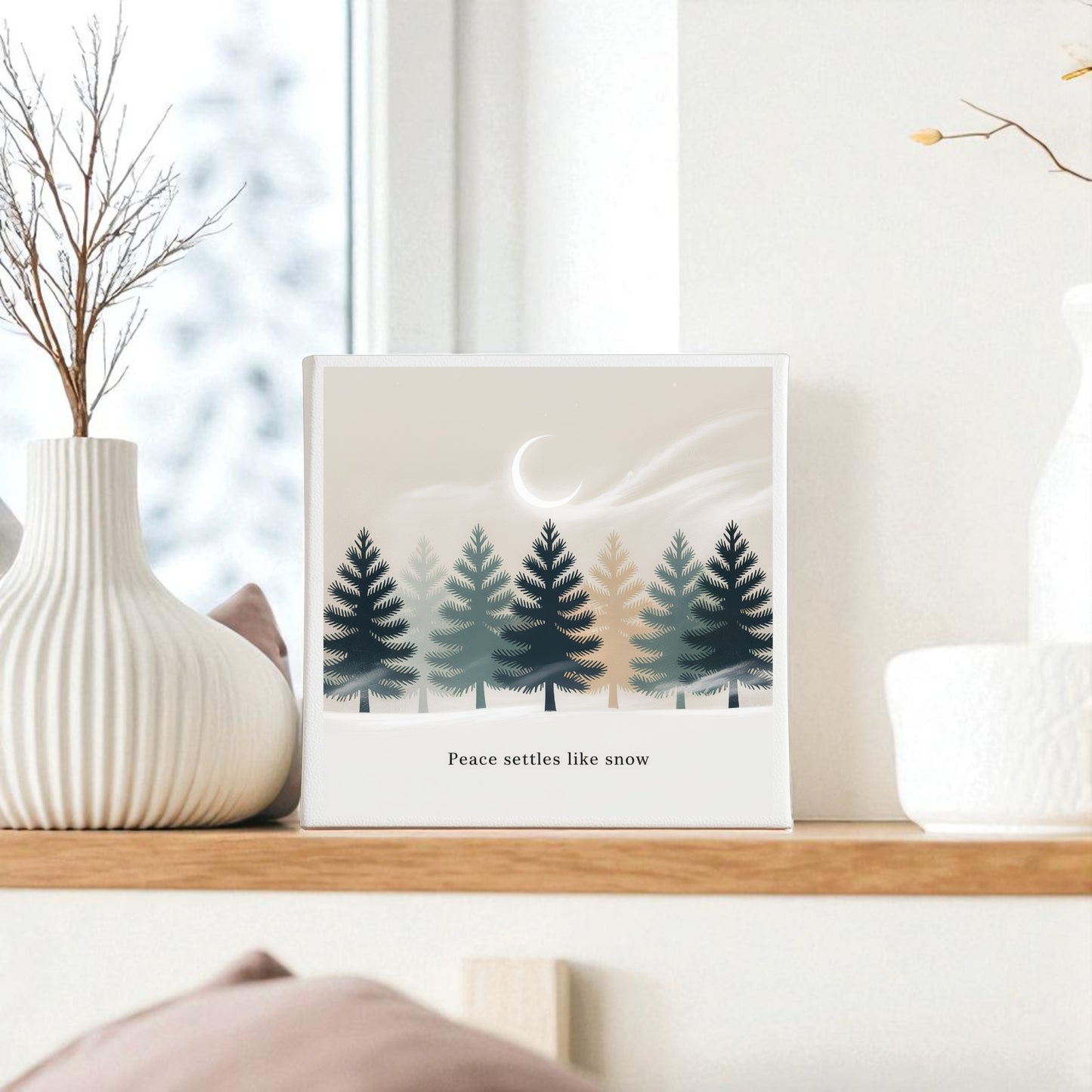 The Peace Settles Like Snow Canvas by Mirabilia Boutique, featuring minimalist pine trees, a crescent moon, and the phrase "Peace settles like snow," is displayed on a wooden shelf beside textured white vases with dried branches.