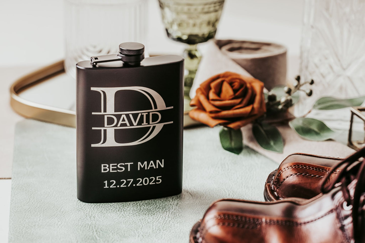 The Personalized Best Man Flask Gift Set by Mirabilia Boutique, engraved with "DAVID," "BEST MAN," and "12.27.2025," sits beside a brown rose, green glass, and dress shoes—a refined custom groomsmen present for your special day.