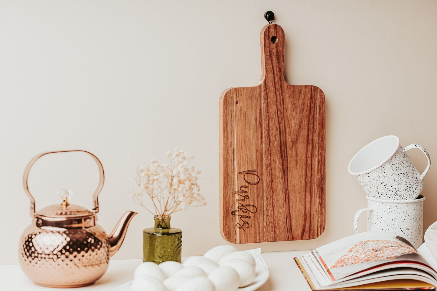 A Mirabilia Boutique Engraved Acacia Cutting Board (14x8), white cups, a copper teapot, eggs on a plate, a small green vase with dried flowers, and an open book sit on a kitchen counter with a light background.