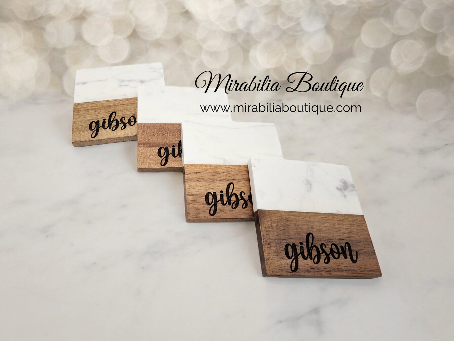 The Personalized Marble and Acacia Coaster Set by Mirabilia Boutique features custom-engraved coasters with white marble tops and wooden bottoms, perfect as a housewarming or wedding gift. Displayed in a row at www.mirabiliaboutique.com.