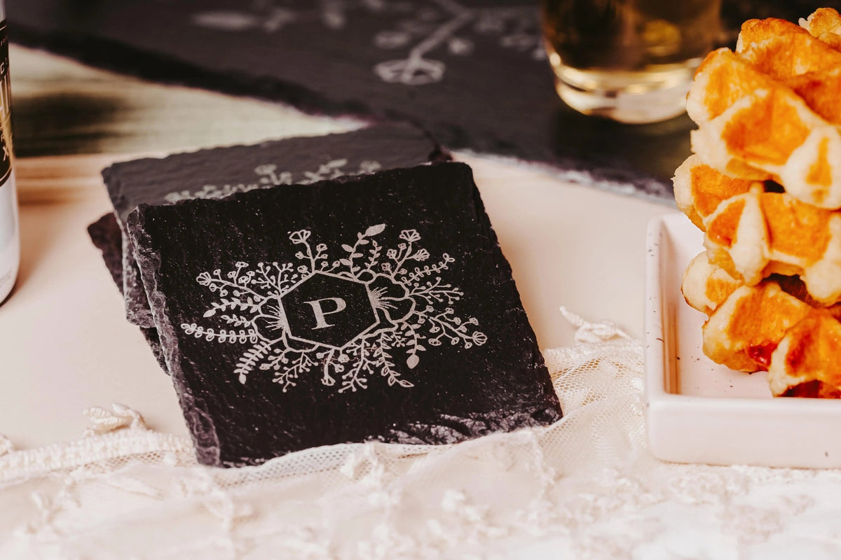A set of four Personalized Slate Coasters by Mirabilia Boutique, engraved with a white floral monogram "P," sits on a lace tablecloth beside waffles—an elegant, custom housewarming gift.