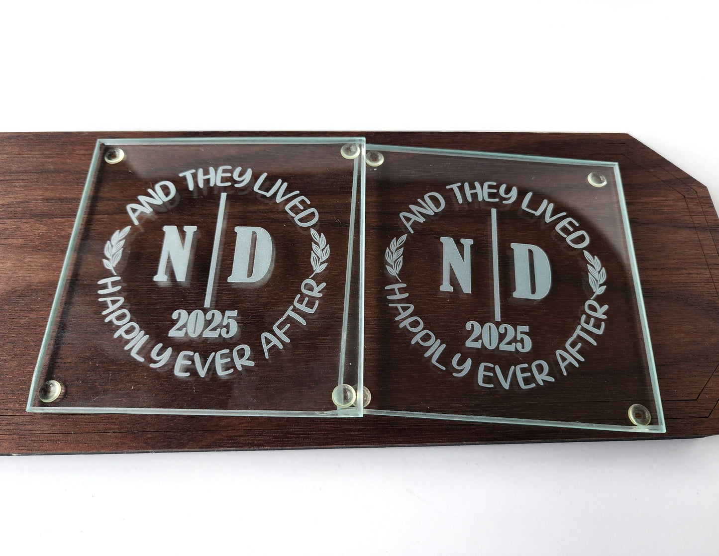 The Personalized Glass Wedding Coasters by Mirabilia Boutique make an ideal wedding gift, featuring custom engraved messages like "AND THEY LIVED HAPPILY EVER AFTER," initials "N D," and the year "2025." Displayed on a wooden base, they add timeless romance to any home décor.