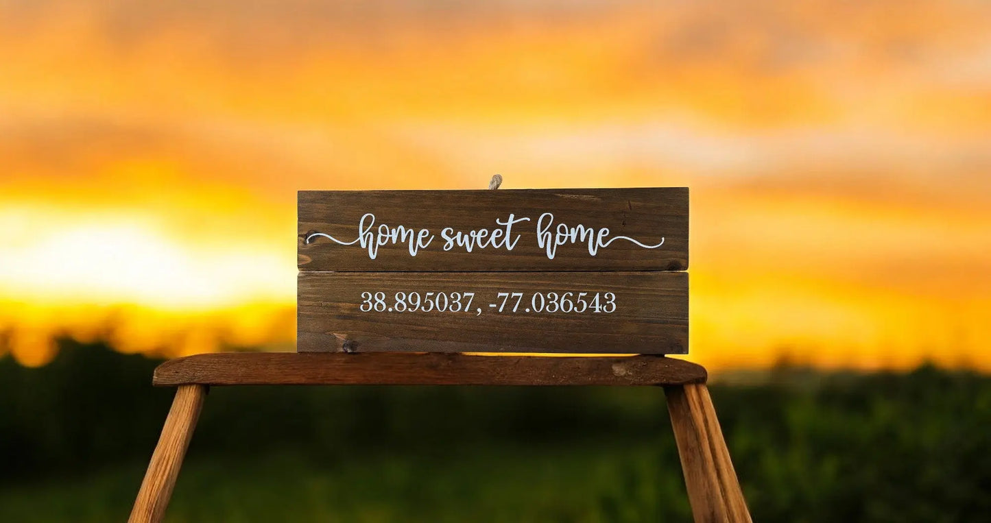 The Mirabilia Boutique Personalized Home Sweet Home Coordinates Wood Sign sits on a stool, featuring custom city longitude and latitude and “home sweet home,” set against a warm orange-yellow sunset background.