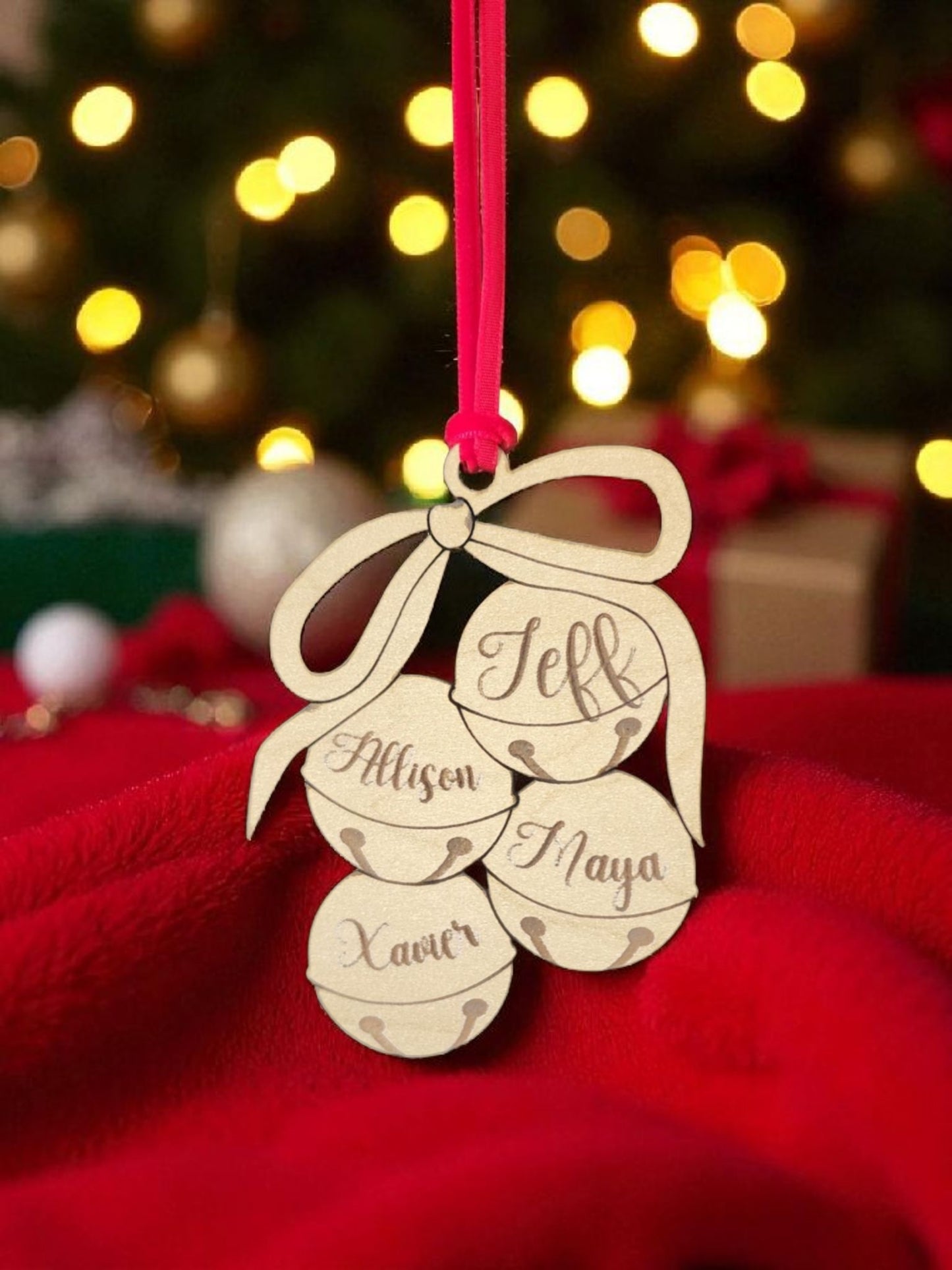 The Custom Family Christmas Ornament by Mirabilia Boutique is a laser-cut wooden holiday tree decoration with four jingle bells personalized with "Allison," "Jeff," "Xaver," and "Naya," hanging by a red ribbon amid festive lights and gifts.