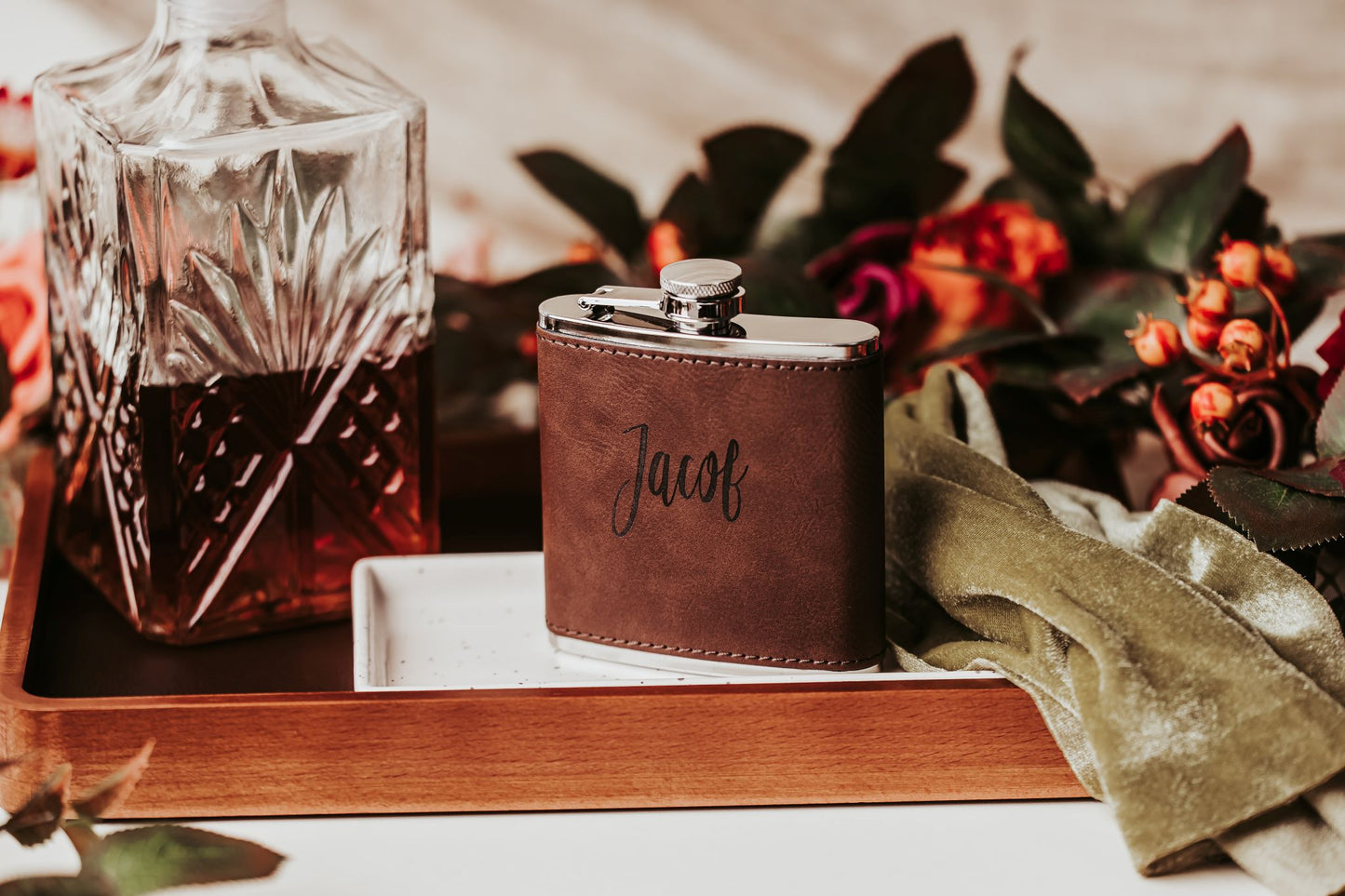 The Custom Engraved Flask by Mirabilia Boutique, personalized with “Jacob,” sits beside a crystal decanter, flowers, and green velvet—an elegant leather gift for him and a perfect groomsmen keepsake.