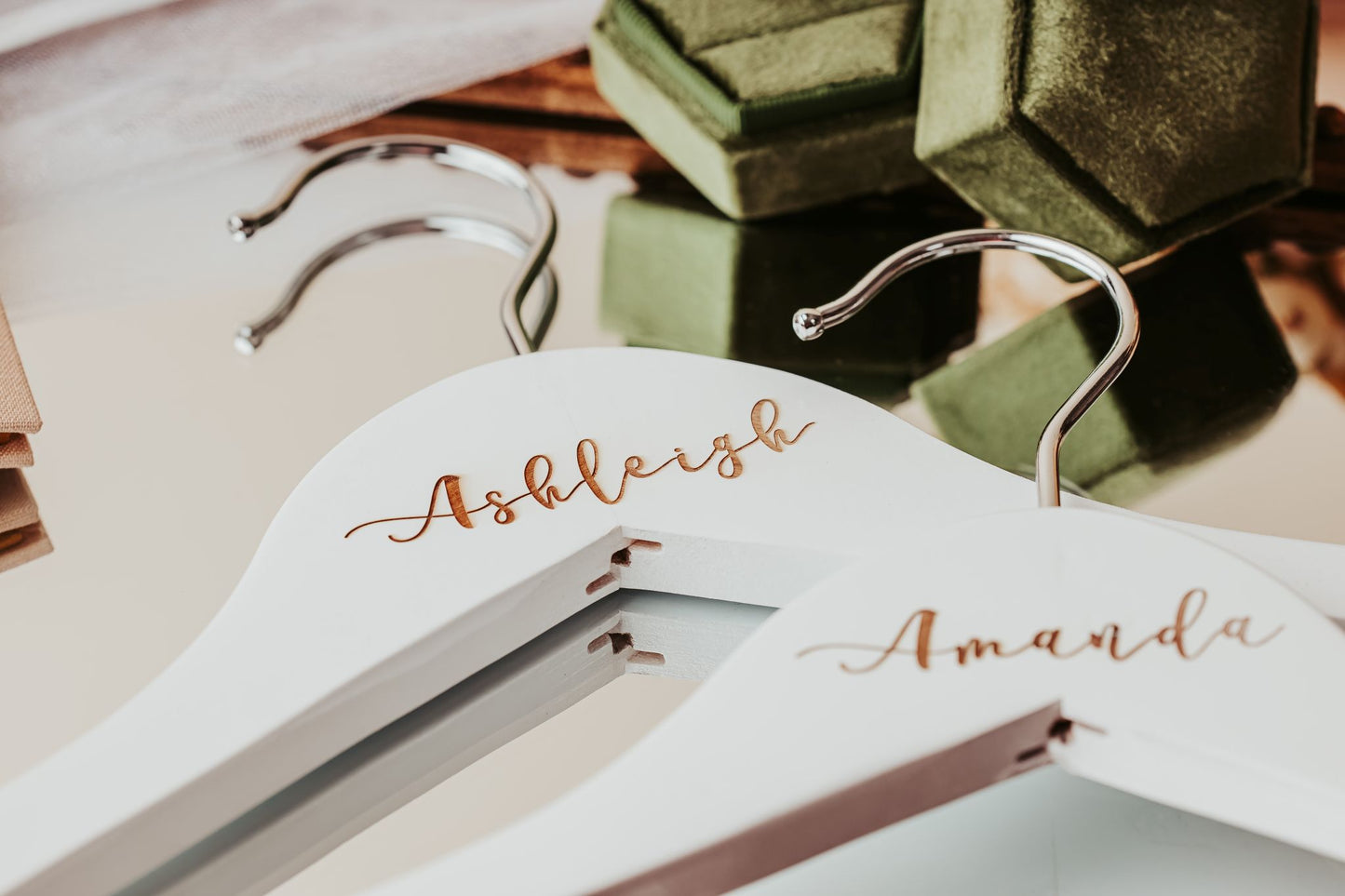 The Personalized Wedding Hangers | Custom Name Hanger Set by Mirabilia Boutique, featuring gold script names, rest on a mirror and add a special touch to wedding day details, with green velvet ring boxes visible in the background.