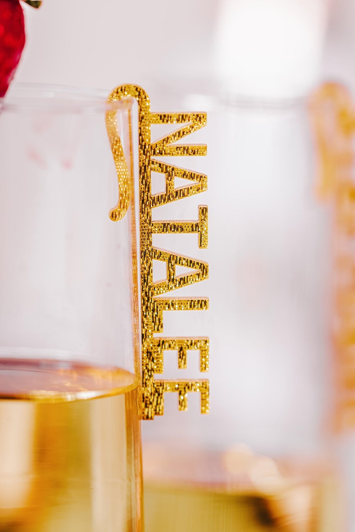 A clear glass with a gold base displays a "NATALIE" custom acrylic wine glass name charm from Mirabilia Boutique, clipped to the rim for stylish personalization; part of a strawberry accents this unique party decor.