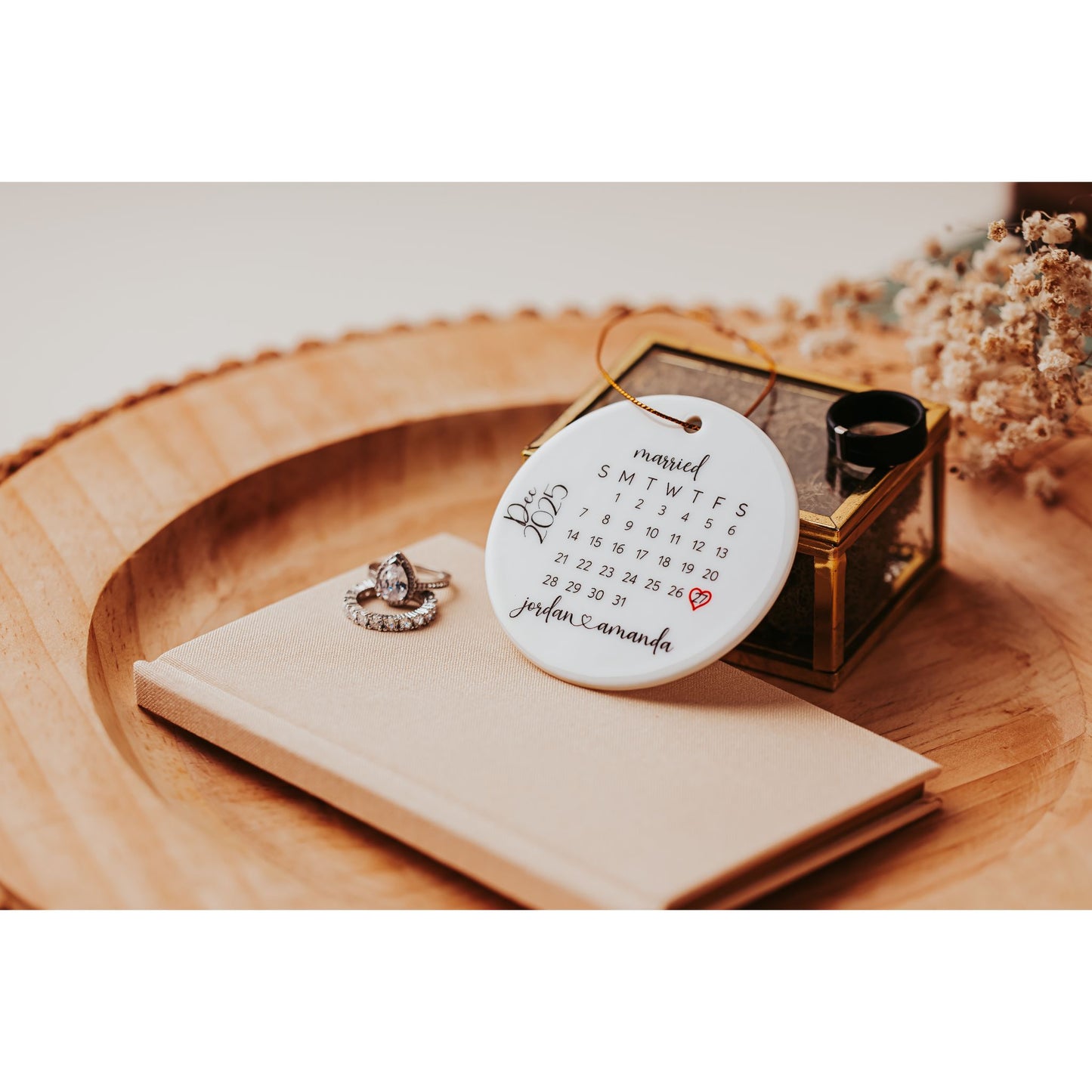 The Personalized Wedding Date Ornament – Custom Ceramic Calendar Keepsake by Mirabilia Boutique rests on a notebook beside an engagement ring and glass box, making a perfect newlyweds keepsake displayed on a wooden tray.
