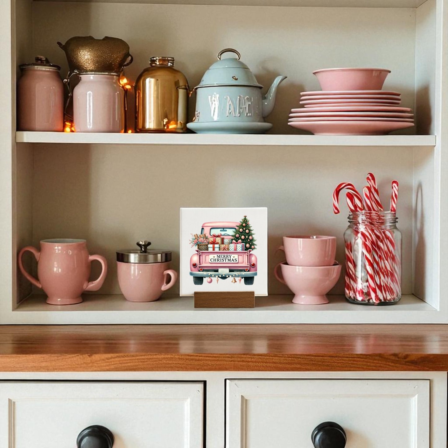 A kitchen shelf with pink dishes, mugs, bowls, a blue teapot, a gold jar, candy cane jar, and the Mirabilia Boutique Pink Christmas Truck Mini Sign—glam holiday decor with gifts & tree for a festive touch.