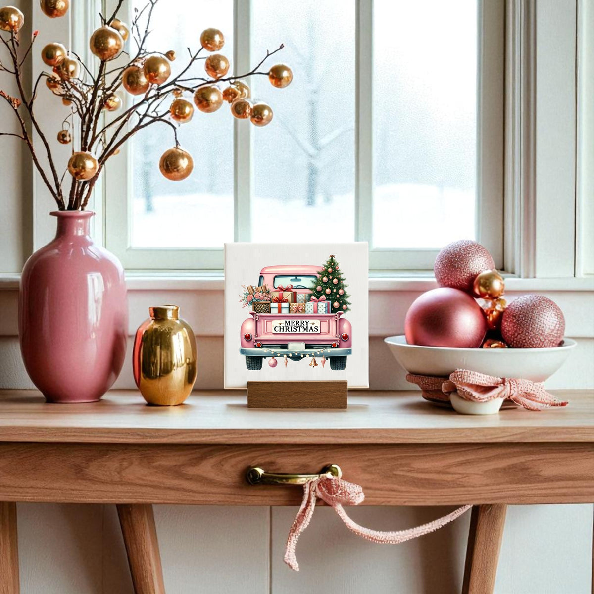 A wooden table decorated with pink and gold ornaments, a pink vase, and a bowl of baubles is topped with the Mirabilia Boutique Pink Christmas Truck Mini Sign. Snow falls outside, completing the glam holiday scene.