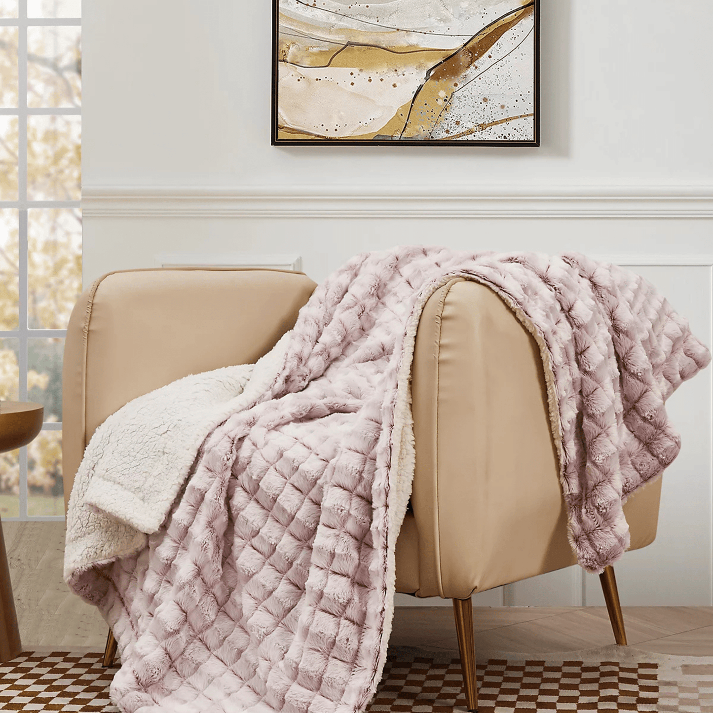 The Custom Fall Throw Blanket by Mirabilia Boutique, featuring a light pink texture and white sherpa lining, is draped over a beige armchair in a modern room with a geometric rug and abstract wall art.