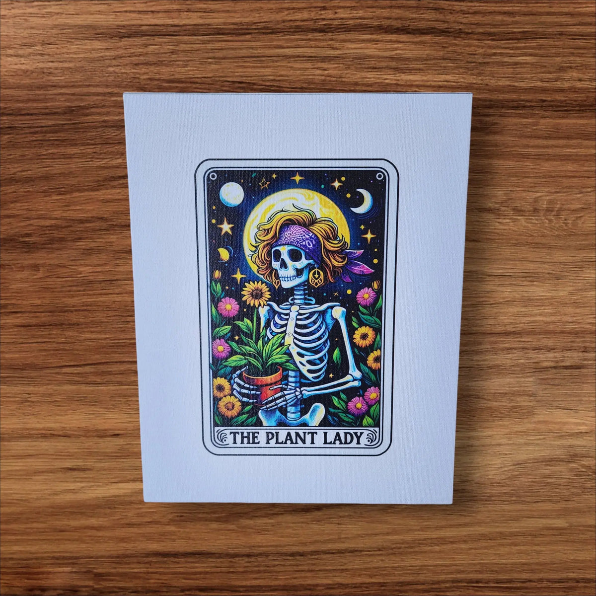 The Plant Lady Canvas Art by Mirabilia Boutique features a stylish skeleton with a purple headscarf and yellow hat, holding a potted plant amid flowers, stars, and a crescent moon. UV printed 8x10—perfect wall decor for plant moms.