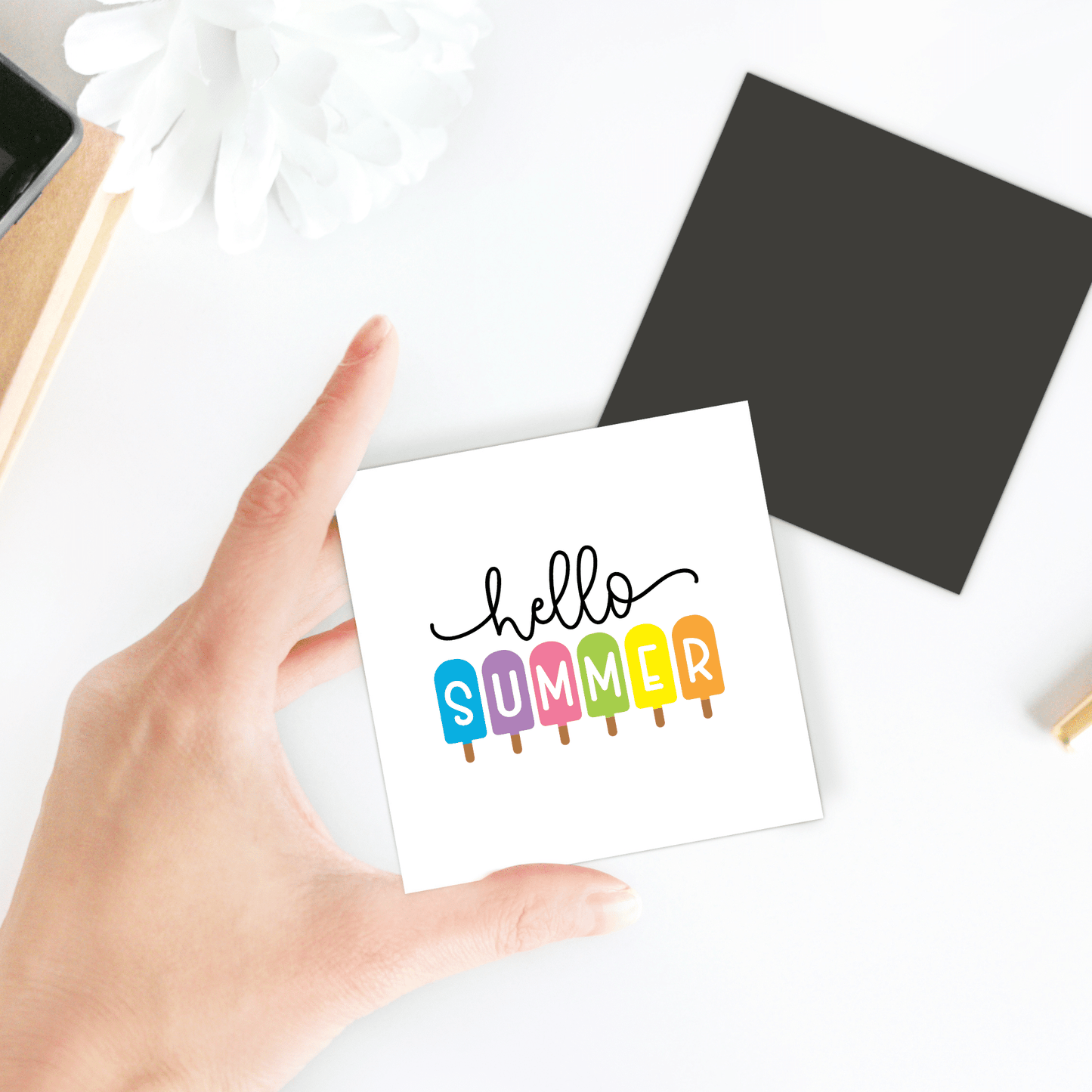 A hand holds the Hello Summer Popsicle Magnet by Mirabilia Boutique, featuring "SUMMER" spelled with rainbow popsicles—a vibrant ice pop fridge art. Nearby: a black square, white flower, and brown object on a white surface.