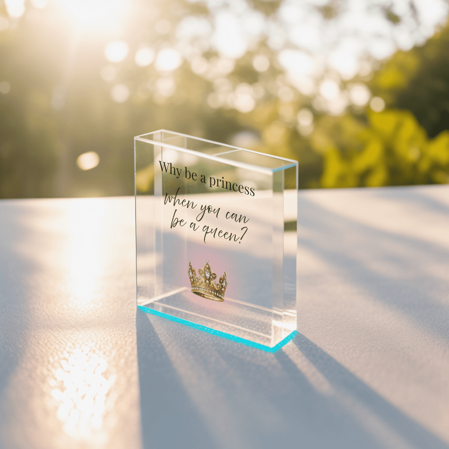 The Mirabilia Boutique "Why Be a Princess When You Can Be a Queen" Acrylic Block Gift for Her features a gold crown design and inspiring message—an empowering decor piece, perfect for gifting, shown on a sunlit surface with blurred greenery behind.