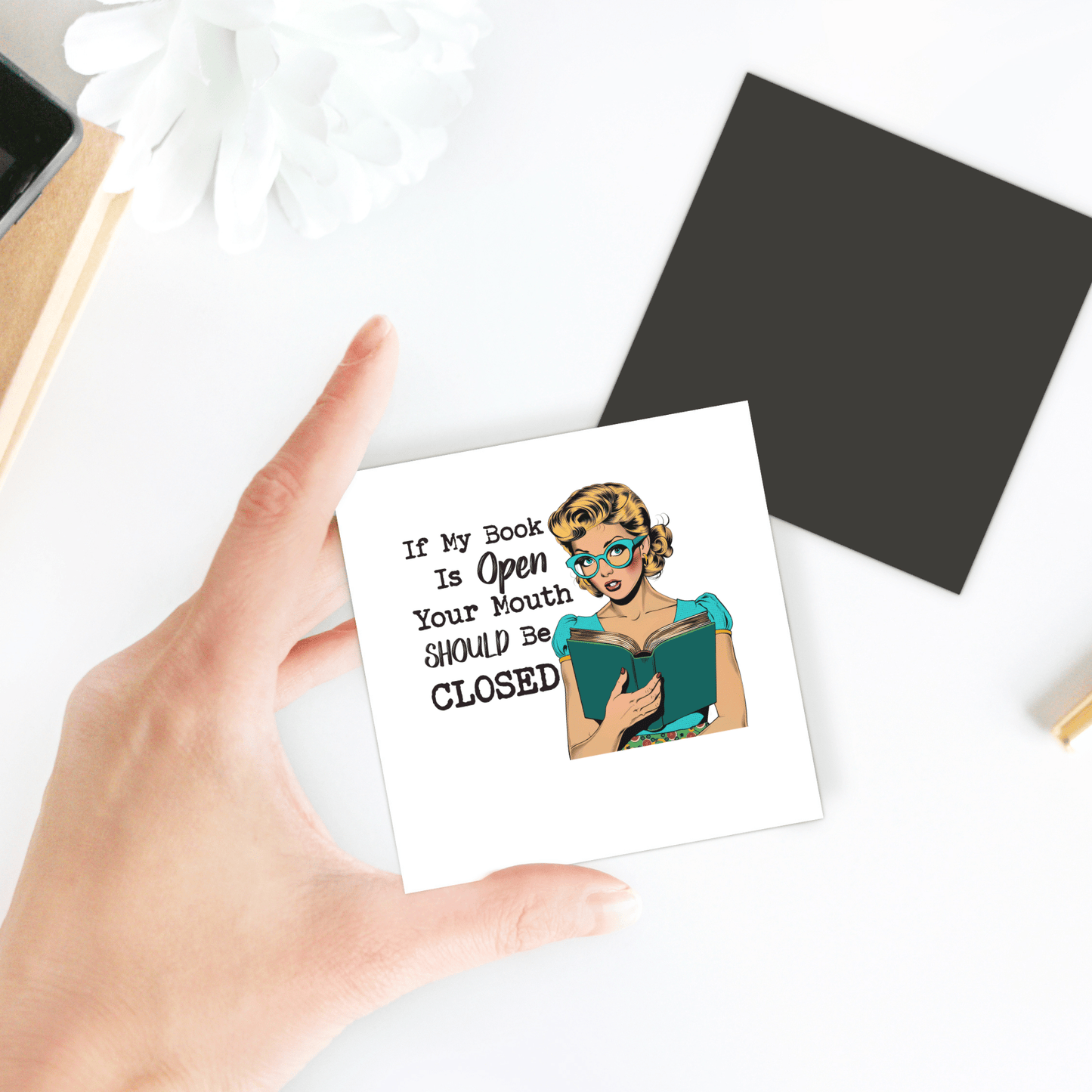 A hand holds the “If My Book Is Open” 3x3 retro magnet by Mirabilia Boutique, a funny UV printed gift for readers and introverts. In the background, a black square and white flower rest on the table.