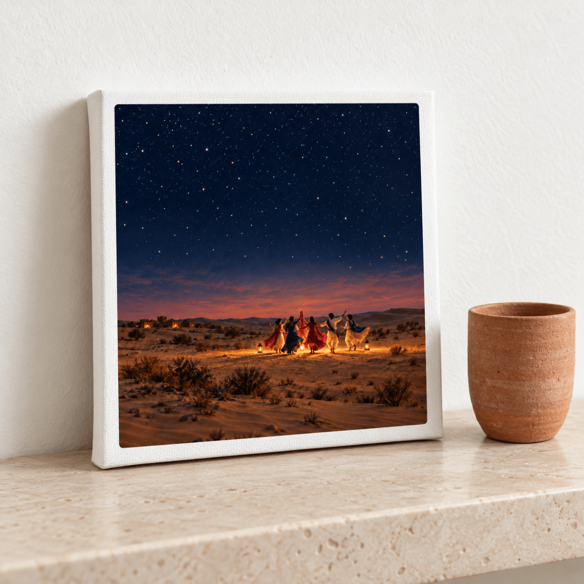 The Night We Danced Canvas | Sand & Saffron by Mirabilia Boutique, a gallery-wrapped canvas depicting people dancing under a starry desert sky, leans against a white wall next to a terracotta pot on a stone ledge.