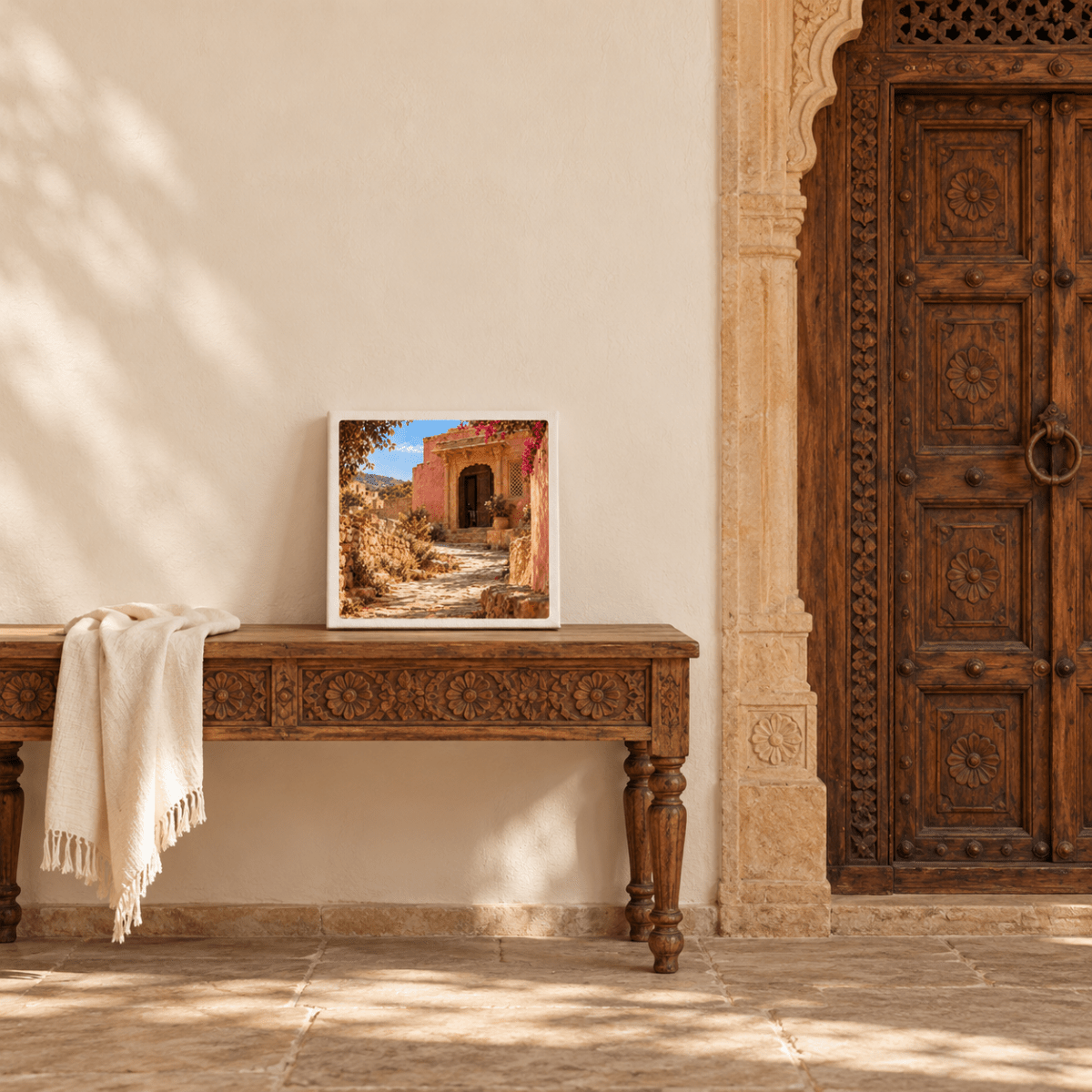 A framed square The House Ahead Canvas | Sand & Saffron print of a sunlit Mediterranean rose-toned house, displayed on a wooden console table next to an ornate carved door.