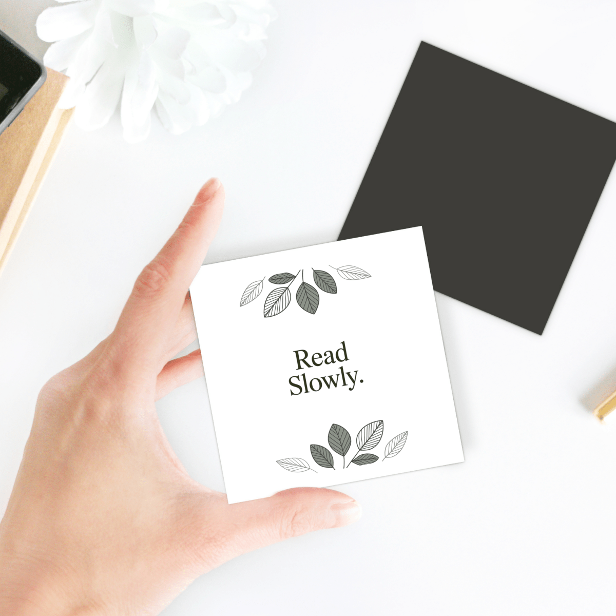A hand holds the Read Slowly Book Lover Magnet by Mirabilia Boutique, a charming gift for readers. In the background, a black square, a white flower, and part of a pen rest on the white surface.