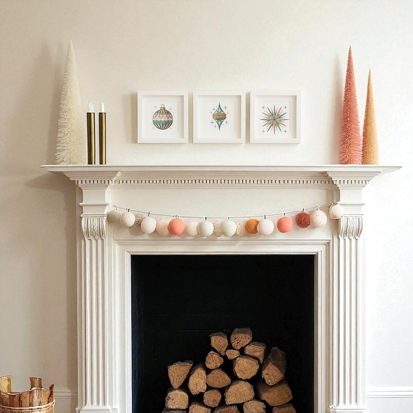 A white mantel displays vintage Christmas decor—cone trees, gold candles, a garland of round ornaments, and Mirabilia Boutique’s Retro Ornament Trio | Mid-Century Holiday Wall Art Set above. Firewood is neatly stacked inside the fireplace.