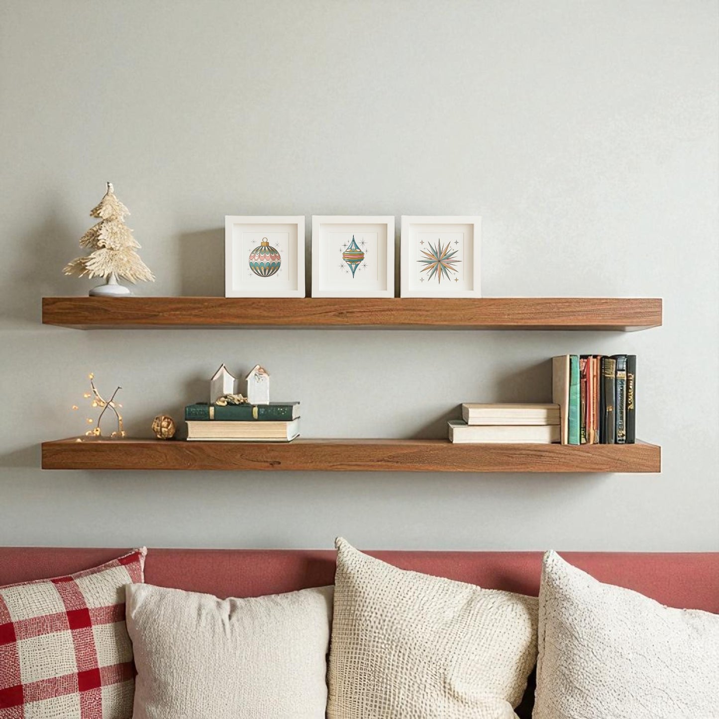 Two wooden floating shelves on a light wall showcase small framed art, books, a miniature tree, and the Retro Ornament Trio Mid-Century Holiday Wall Art Set by Mirabilia Boutique above a sofa with white and red cushions.