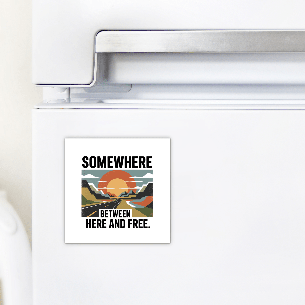 The Somewhere Between Here and Free Magnet from Mirabilia Boutique features a stylized sunset over mountains with the phrase “Somewhere between here and free,” shown here on a white refrigerator.