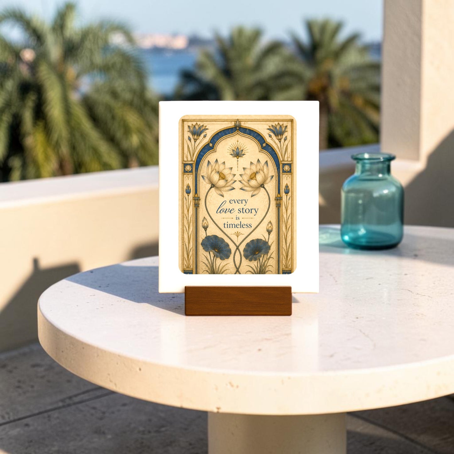 The Mirabilia Boutique “Every Love Story Is Timeless | Lotus Mini Sign,” featuring delicate floral artwork and the heartfelt phrase “every love story is timeless,” rests on a round white table outdoors, set against a backdrop of palm trees and shimmering water.