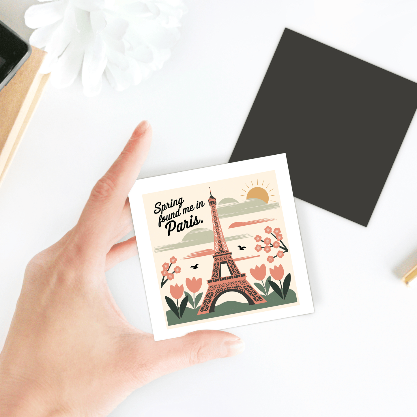 A hand holds the "Spring Found Me in Paris Magnet – Eiffel Tower Charm" by Mirabilia Boutique, featuring flowers, birds, and the sun. A black square and flower sit on a white table, evoking a charming spring keepsake.