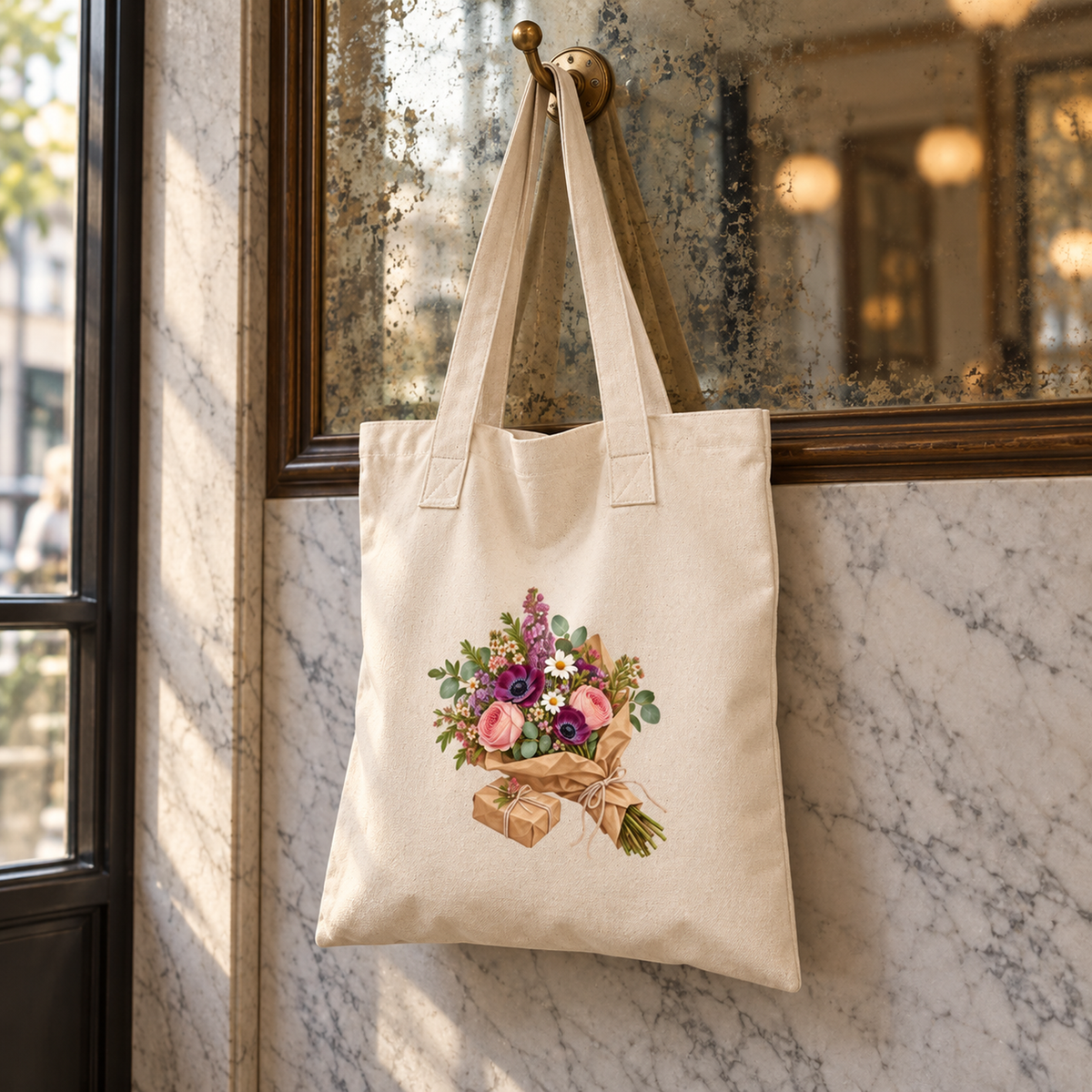 Market Morning Tote Bag by Mirabilia Boutique, a natural canvas bag printed with a floral bouquet and a small gift box, hanging from a hook on a marble wall.