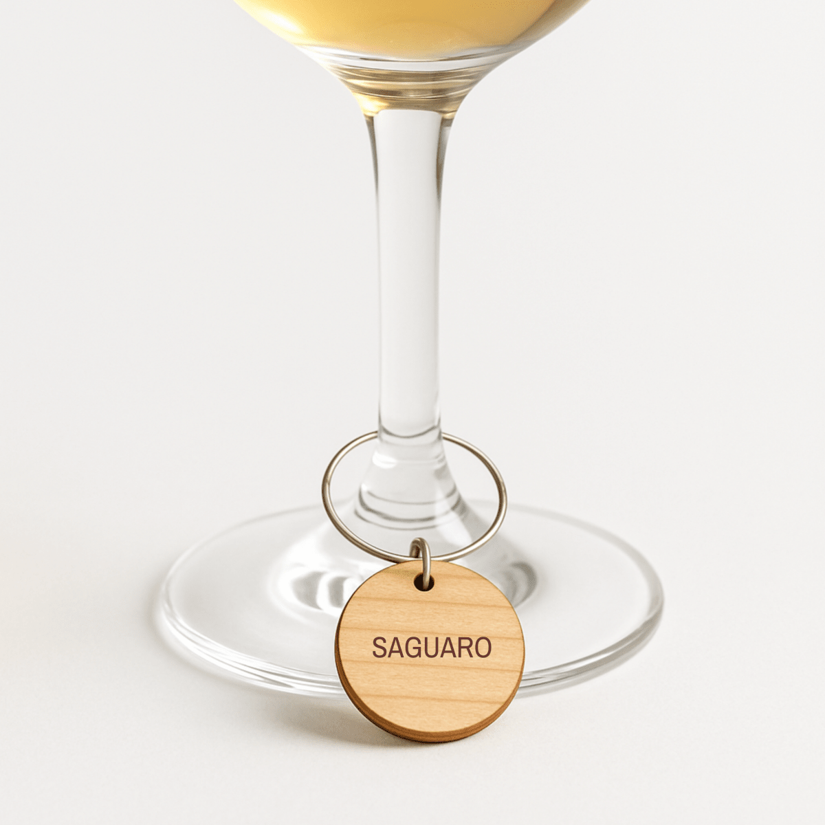 A clear wine glass filled with white wine is adorned with a Desert Dreaming Wine Charm from Mirabilia Boutique, featuring a wooden "SAGUARO" label, shown against a plain white background.
