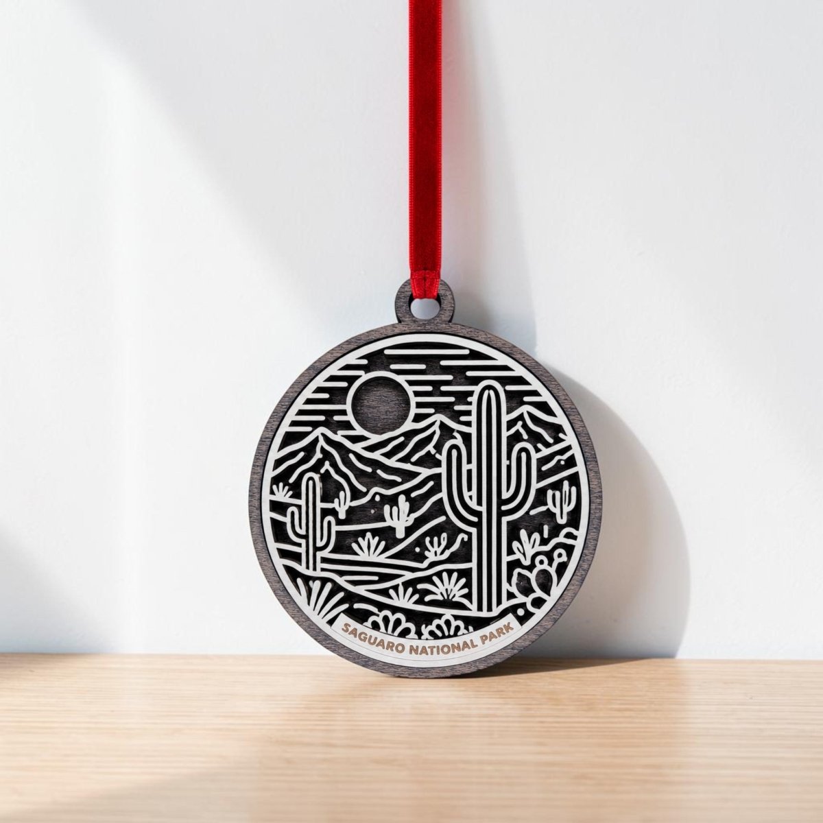 The Saguaro National Park Line Art Christmas Ornament by Mirabilia Boutique features a round design with a red ribbon and an engraved desert landscape of cacti, mountains, and a sun or moon. This ornament is a stunning keepsake for desert lovers, with “Saguaro National Park” inscribed at the base, and is pictured resting against a white wall on a wooden surface.
